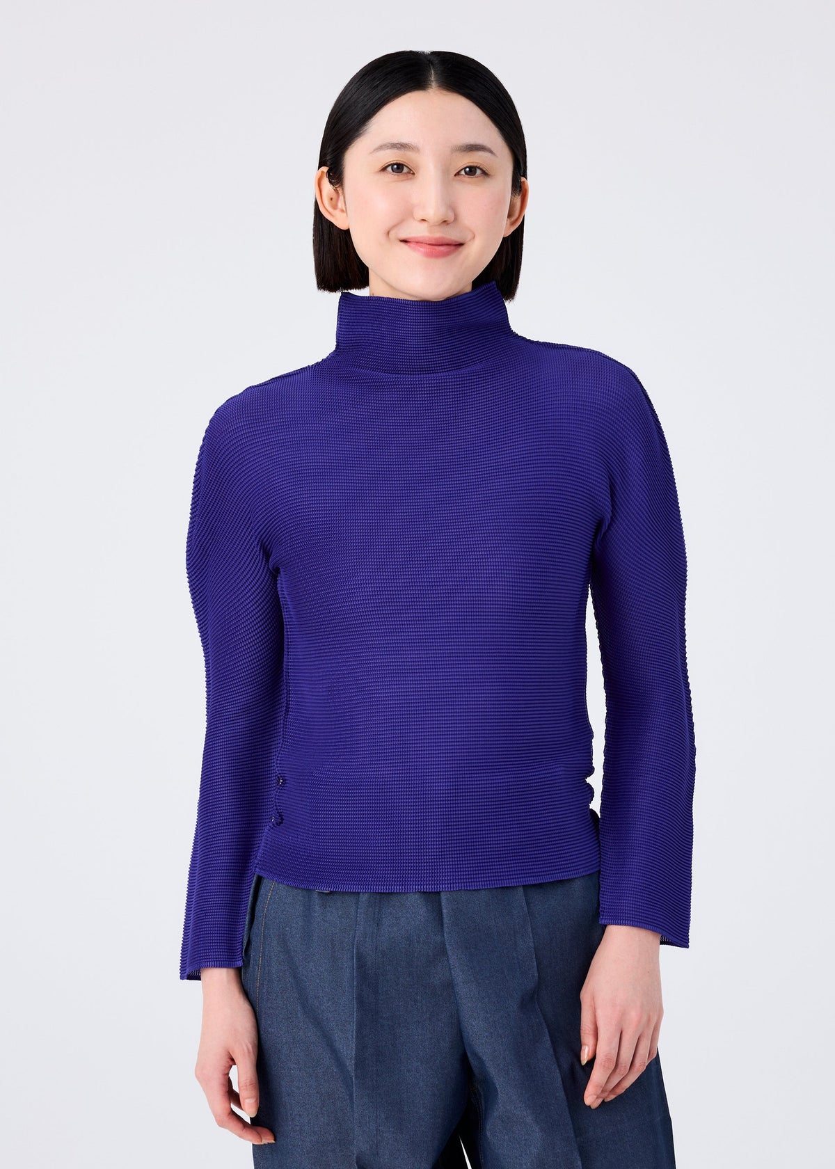 A person with straight dark hair smiles softly against a plain light background, wearing the STRETCH PLEATS 1 royal blue long-sleeved high-neck top and dark blue pants by me ISSEY MIYAKE. view 5 of 7