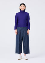 A person stands against a plain white background, smiling and facing forward with arms relaxed, wearing the me ISSEY MIYAKE STRETCH PLEATS 1 purple long-sleeved high-neck top, wide-leg blue denim pants, and white slip-on shoes.