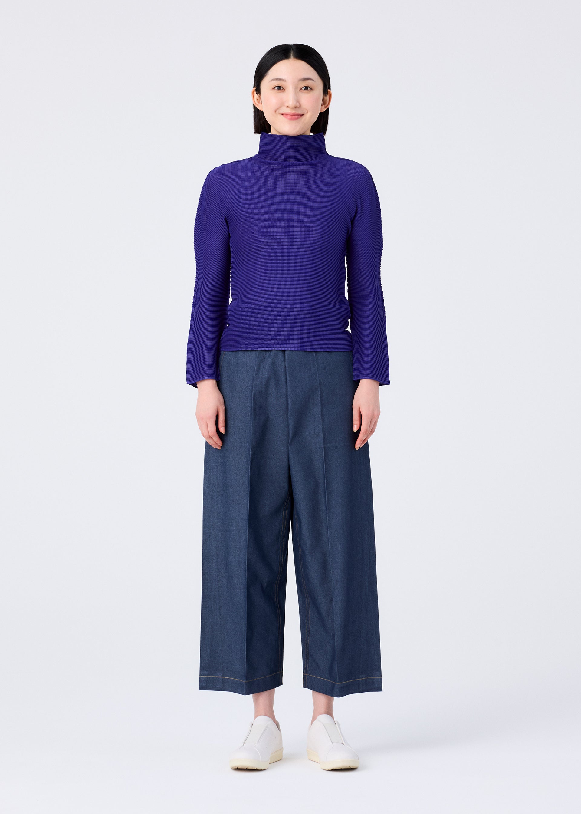 Zoomed view A person stands against a plain white background, smiling and facing forward with arms relaxed, wearing the me ISSEY MIYAKE STRETCH PLEATS 1 purple long-sleeved high-neck top, wide-leg blue denim pants, and white slip-on shoes.