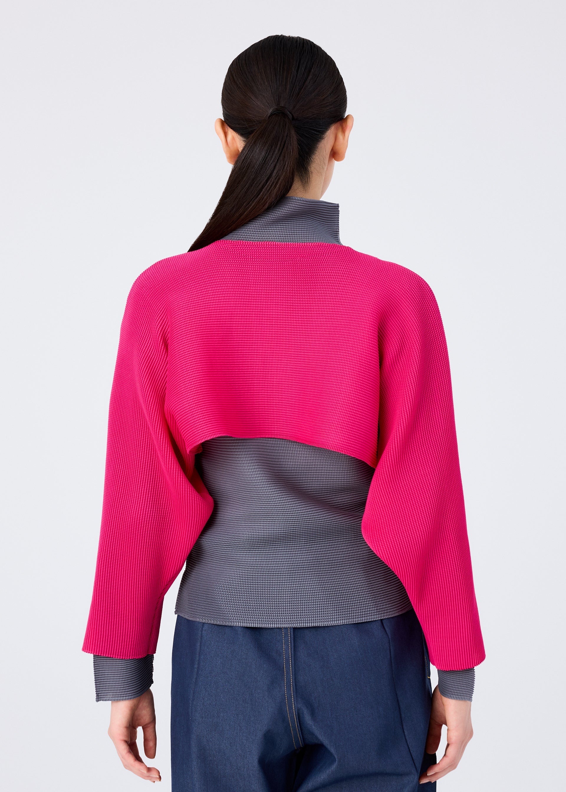Zoomed view A person stands with their back to the camera, wearing blue pants and the me ISSEY MIYAKE DOLMAN STRETCH PLEATS 1 sweater—bright pink, cropped, with dolman sleeves—over a gray turtleneck. Their dark hair is in a low ponytail.