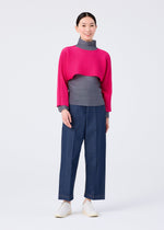 A woman smiles in the me ISSEY MIYAKE DOLMAN STRETCH PLEATS 1: a bright pink crop top sweater over a gray turtleneck with dolman sleeves, wide-leg blue pants, and white shoes, posed against a plain white background.