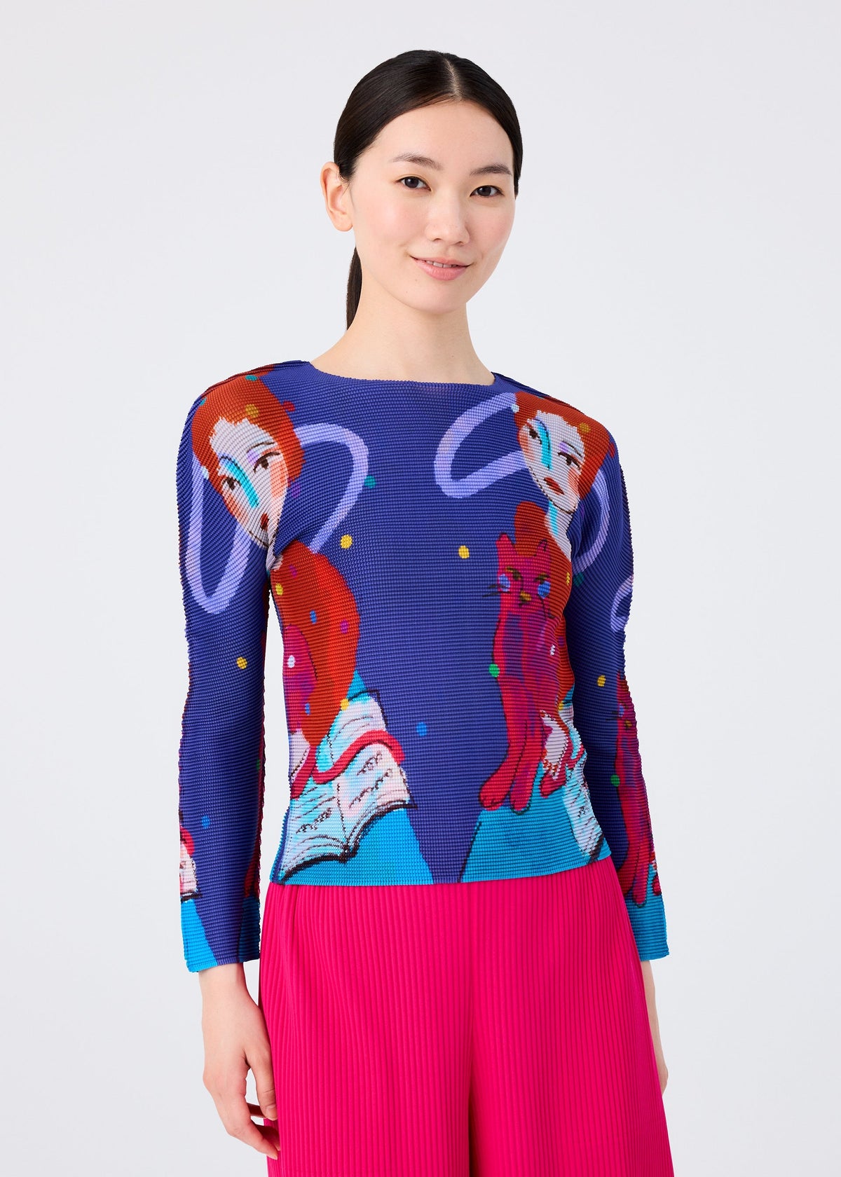 A woman with straight black hair wears the me ISSEY MIYAKE CAT sweater, featuring red-haired figures and books in a wizard print style. She pairs it with bright pink pleated pants and smiles slightly against a light backdrop. view 5 of 7