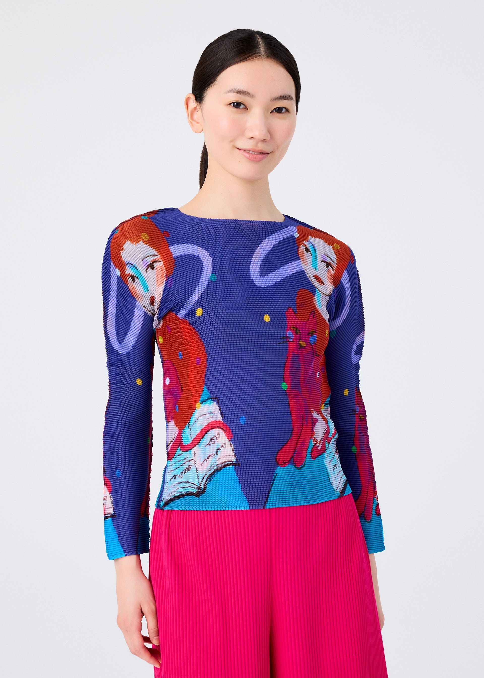 Zoomed view A woman with straight black hair wears the me ISSEY MIYAKE CAT sweater, featuring red-haired figures and books in a wizard print style. She pairs it with bright pink pleated pants and smiles slightly against a light backdrop.