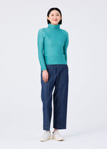 A person stands smiling in a teal long-sleeved STRETCH PLEATS 2 top and loose blue pants by me ISSEY MIYAKE, paired with white shoes against a plain white background.