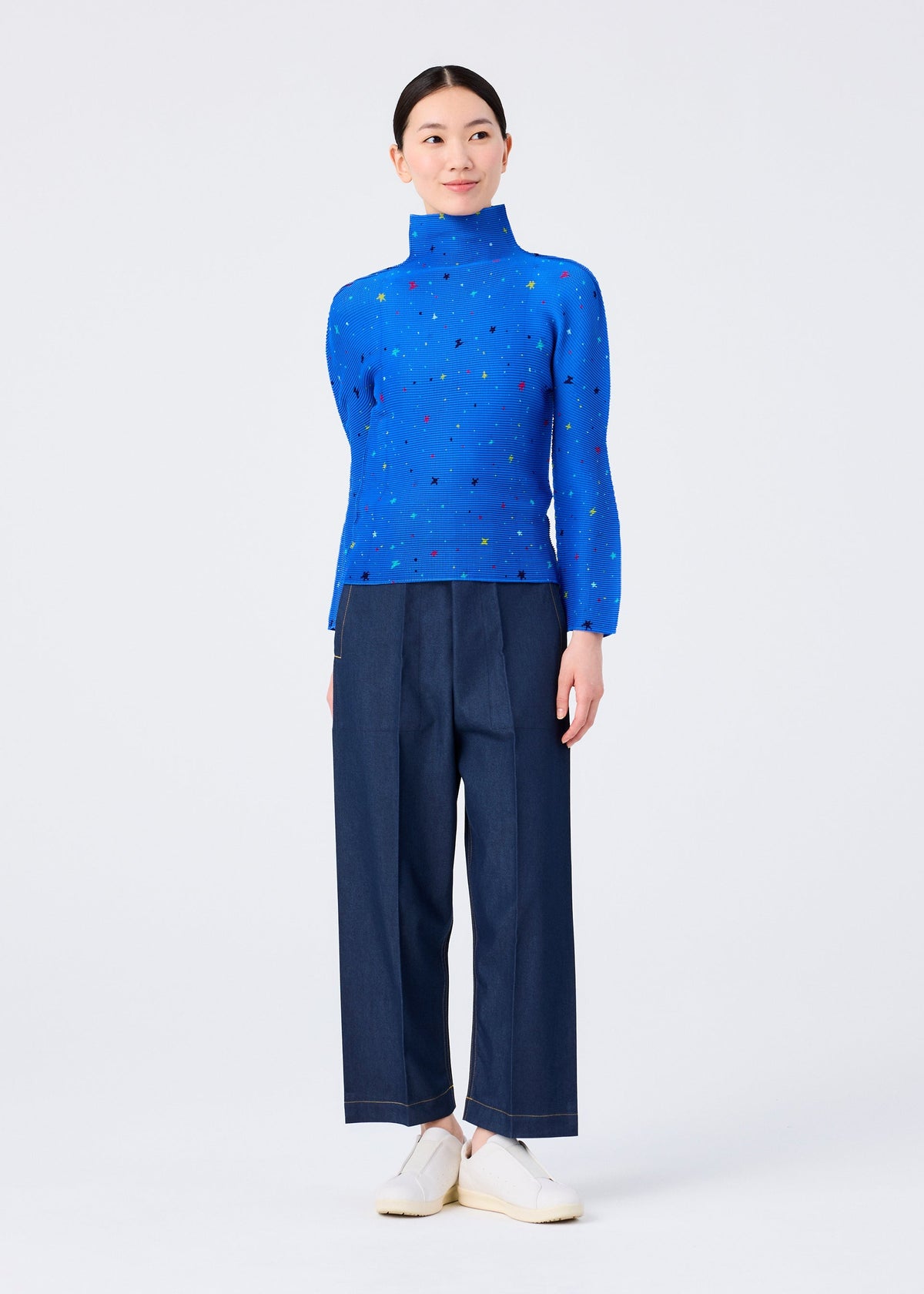 Wearing the TWINKLE STAR high-neck top by me ISSEY MIYAKE with a colorful star print, dark blue wide-leg pants, and white slip-on shoes, the person stands against a plain white background, smiling softly and looking slightly to the side. view 6 of 6