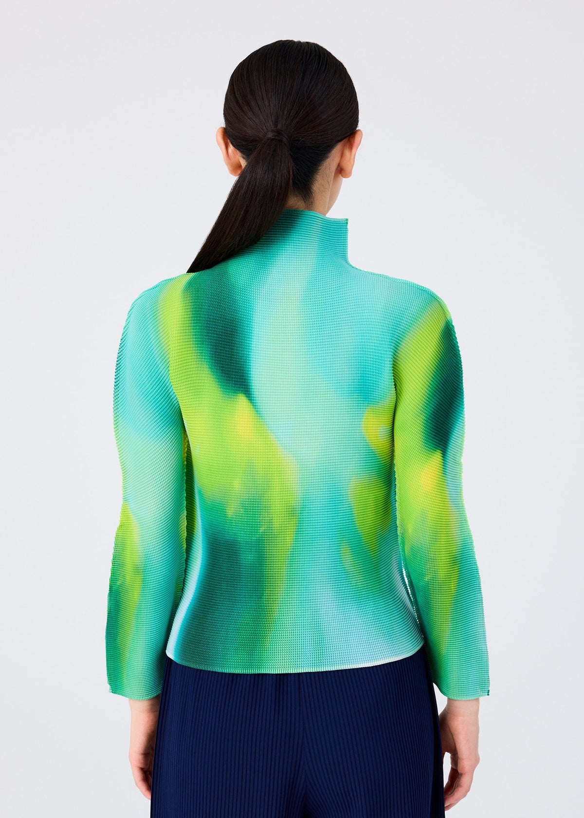 A person with long dark hair in a low ponytail is seen from behind, wearing the me ISSEY MIYAKE AURORA fitted high-neck, long-sleeved top with a green and yellow abstract pattern and navy pleated pants. view 5 of 6