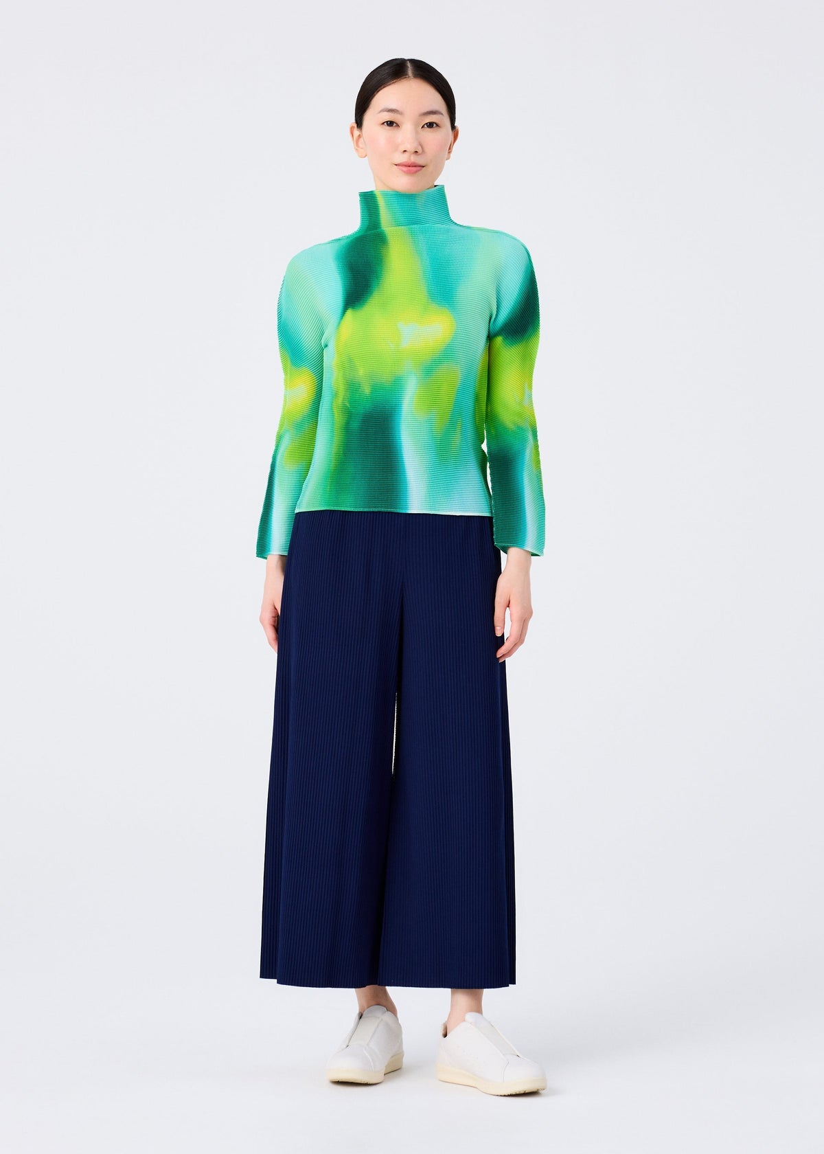 A woman stands facing forward against a white background, wearing the AURORA high-neck long-sleeved top by me ISSEY MIYAKE in green and yellow, paired with navy wide-leg pants and white slip-on shoes. view 6 of 6
