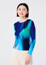 A woman with short dark hair wears the AURORA round-neck long-sleeve top by me ISSEY MIYAKE, featuring a blue, green, and black aurora pattern, paired with light pleated pants. She stands against a plain background, smiling slightly.