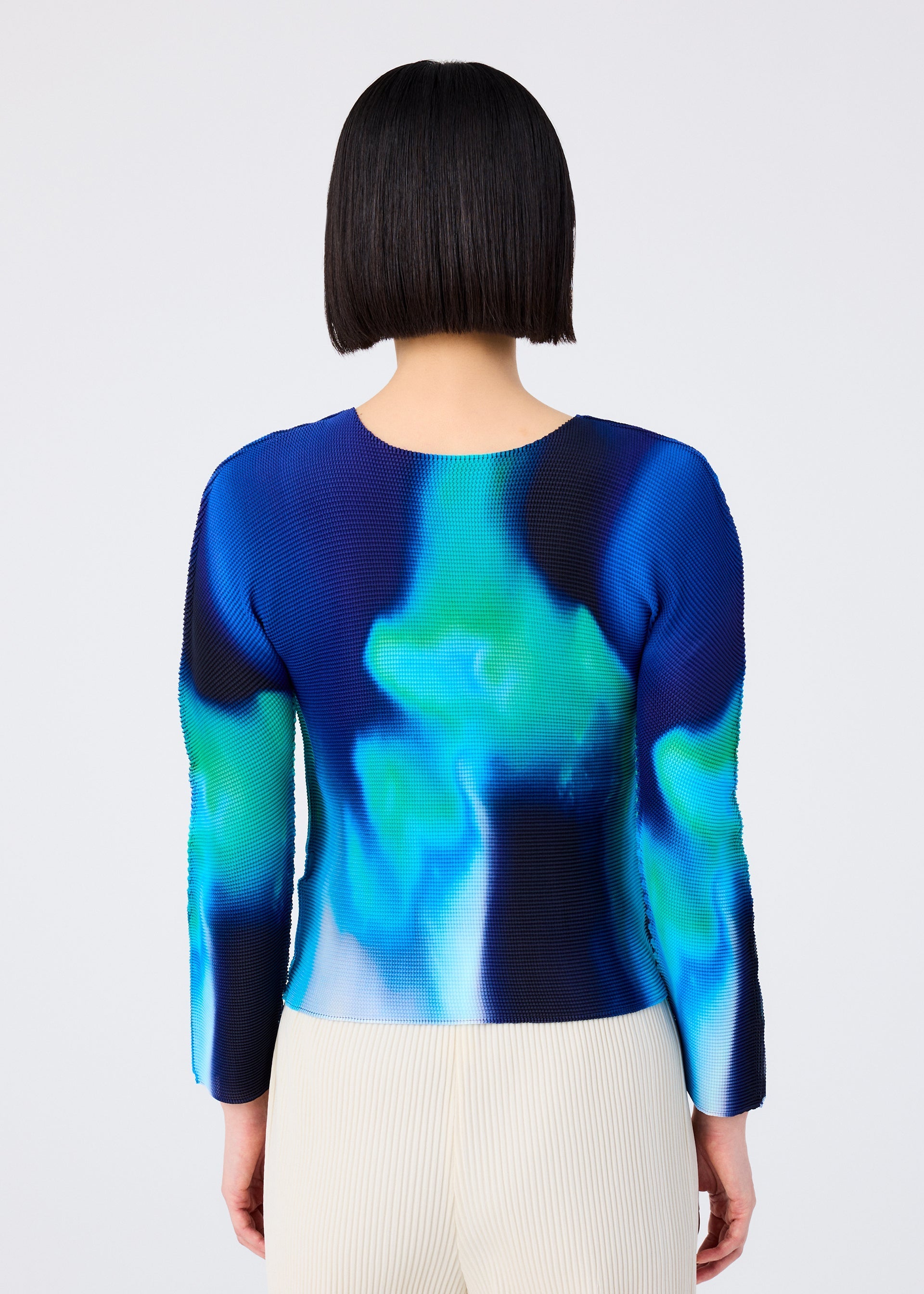 Zoomed view A person with a straight bob is shown from the back, wearing the me ISSEY MIYAKE AURORA round-neck long-sleeve top in blue-green aurora pattern, paired with light pleated pants against a plain background.