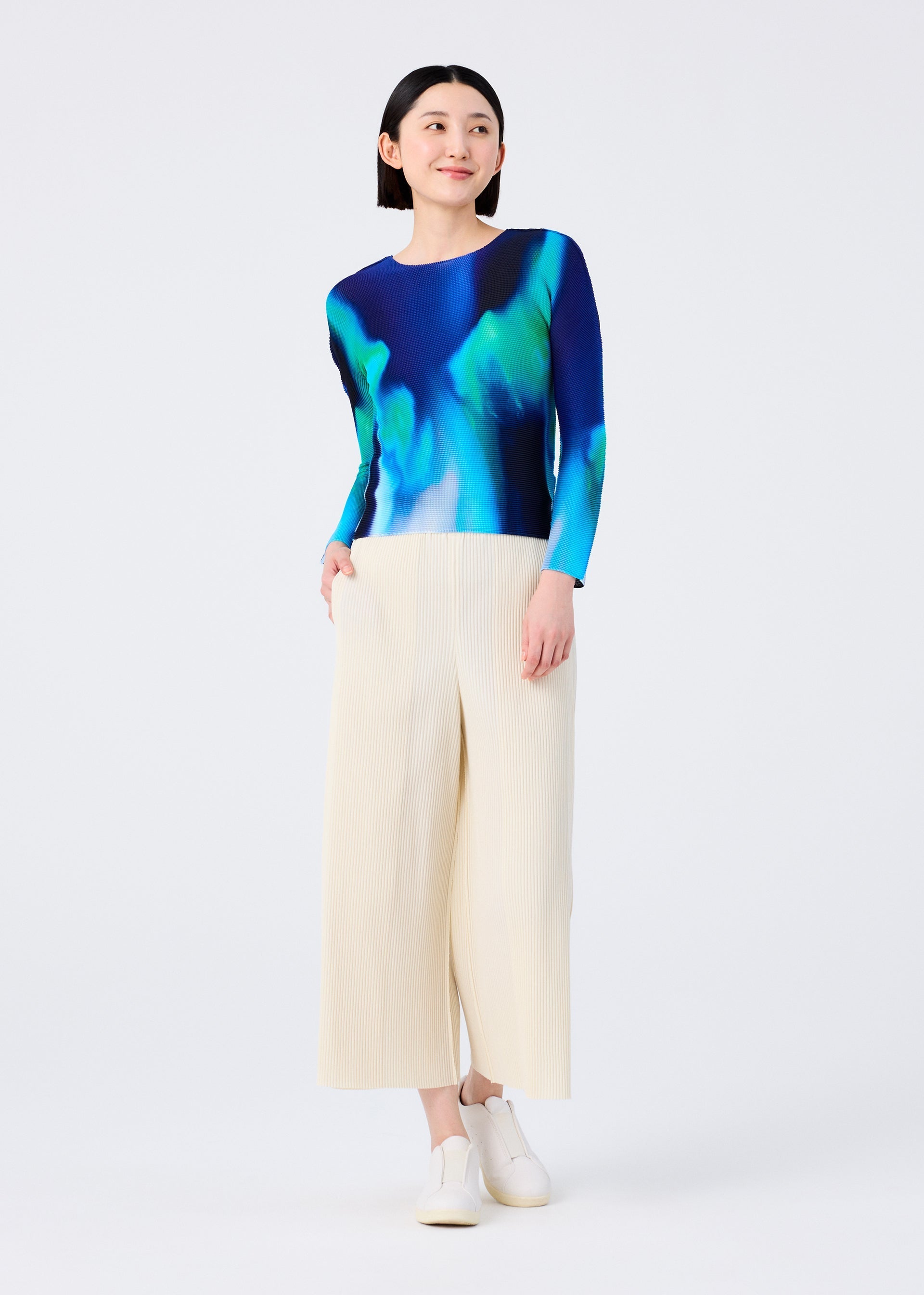 Zoomed view A woman smiles in the AURORA round-neck long-sleeve top by me ISSEY MIYAKE, featuring blue and green aurora patterns, paired with wide-leg cream pleated trousers and white slip-on shoes against a plain white background.