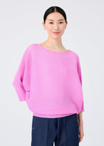 A person with dark hair pulled back wears the me ISSEY MIYAKE DOLMAN STRETCH PLEATS 2 top in light pink with wide 3/4 sleeves and blue pants, standing against a plain light gray background and smiling softly.