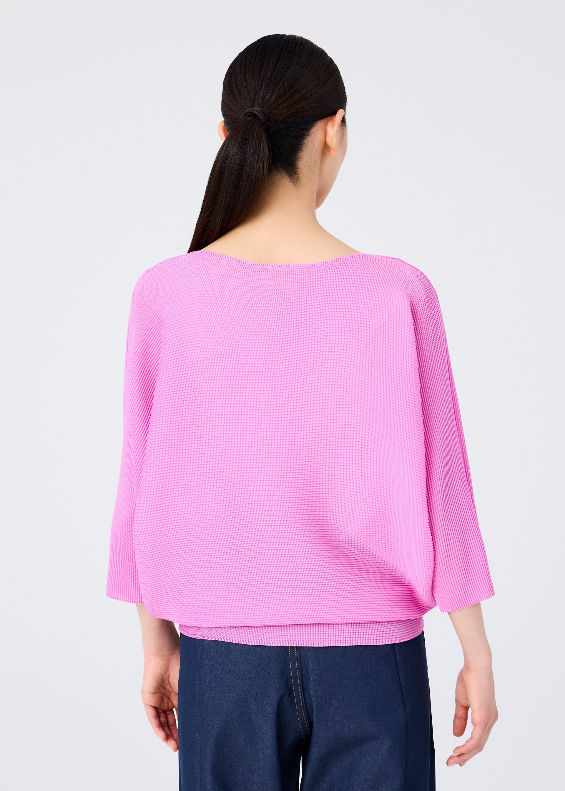 Zoomed view A person with long dark hair in a low ponytail is shown from the back, wearing a loose light pink me ISSEY MIYAKE DOLMAN STRETCH PLEATS 2 top with 3/4 sleeves and dark blue pants, set against a plain light background.