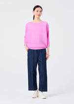 A woman wears the me ISSEY MIYAKE DOLMAN STRETCH PLEATS 2 top in bright pink, paired with navy wide-leg pants and white shoes. She stands against a white background, her straight dark hair pulled back, smiling gently at the camera.
