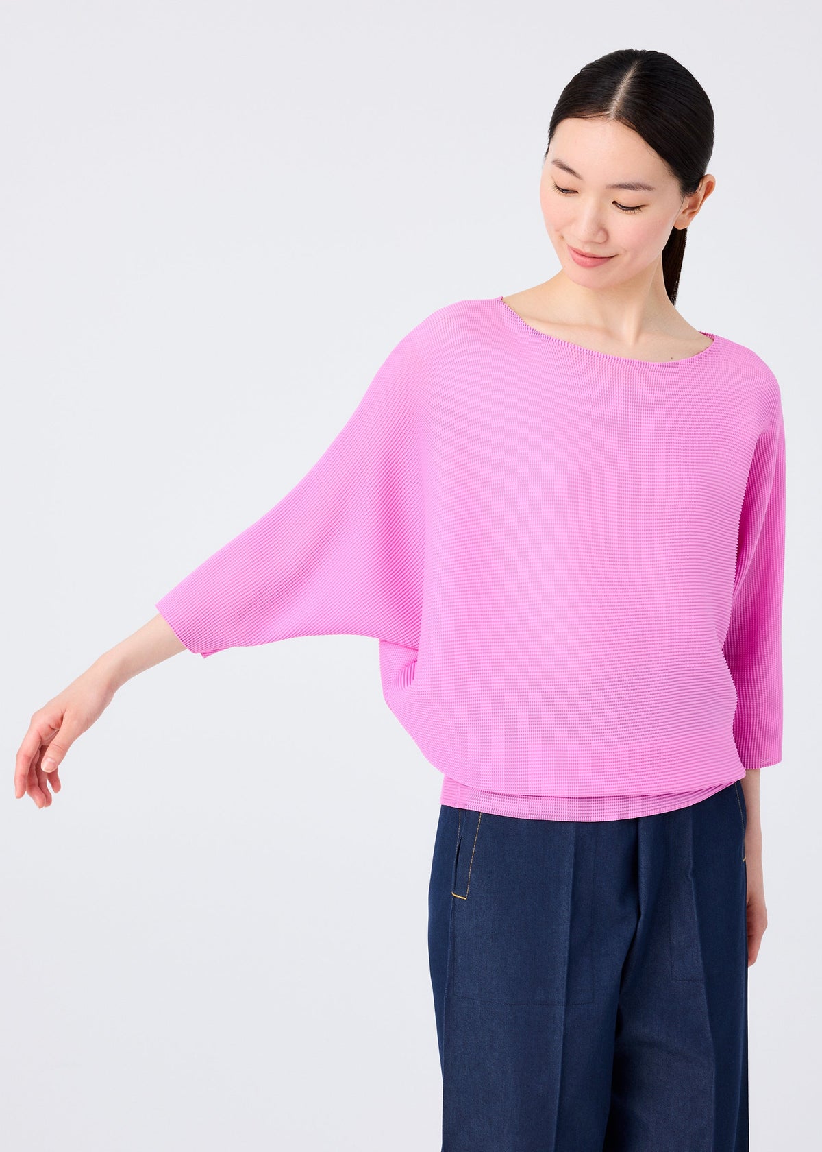 A woman in a loose, light pink DOLMAN STRETCH PLEATS 2 top by me ISSEY MIYAKE and dark blue pants stands against a plain background, smiling and looking down with one arm extended to the side. view 8 of 8