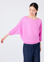 A woman in a loose, light pink DOLMAN STRETCH PLEATS 2 top by me ISSEY MIYAKE and dark blue pants stands against a plain background, smiling and looking down with one arm extended to the side.