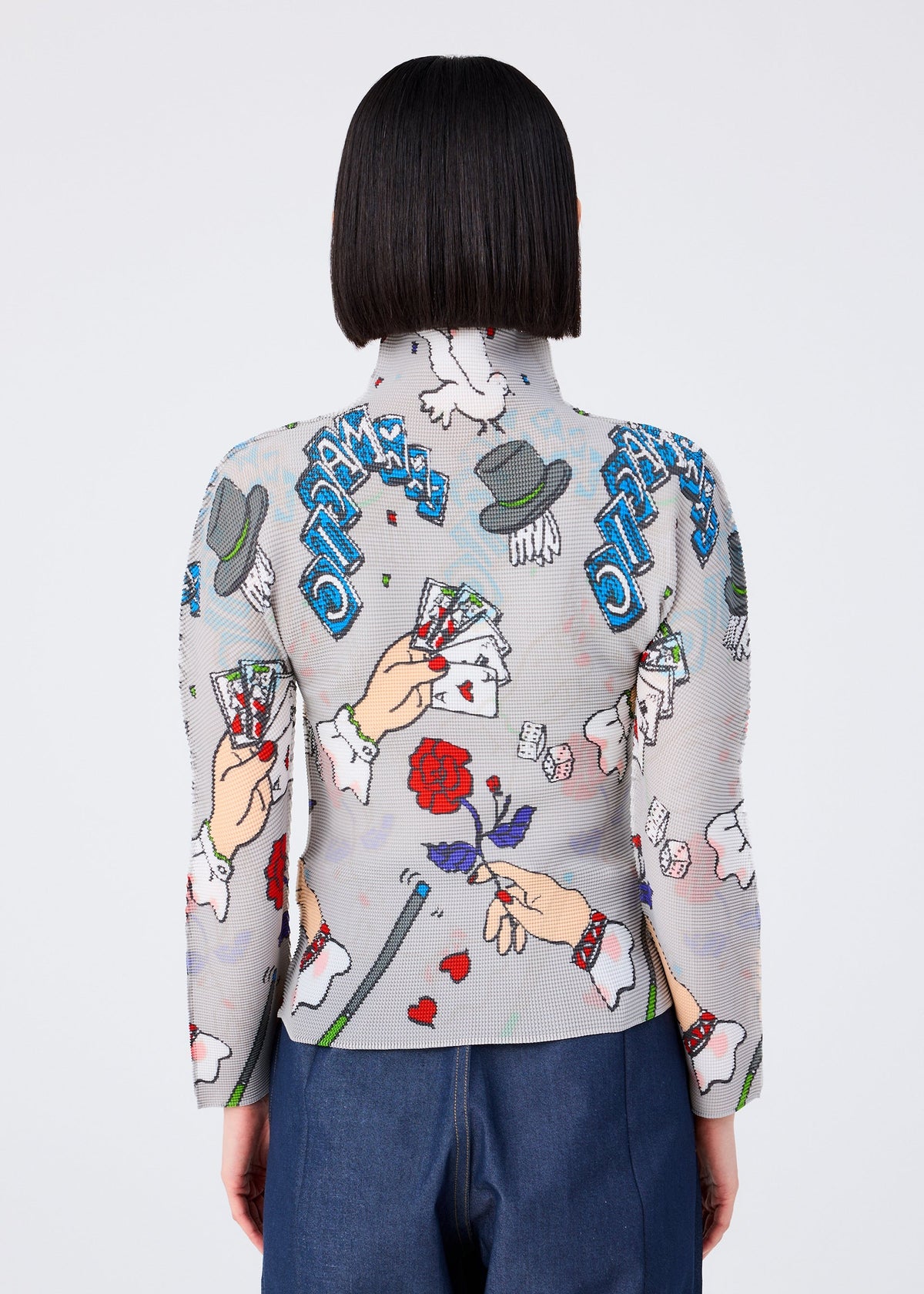 A person with straight, chin-length dark hair faces away, wearing the MAGIC fitted high-neck long-sleeved top by me ISSEY MIYAKE, with colorful cartoon graphics. Denim pants are partially visible. view 5 of 6
