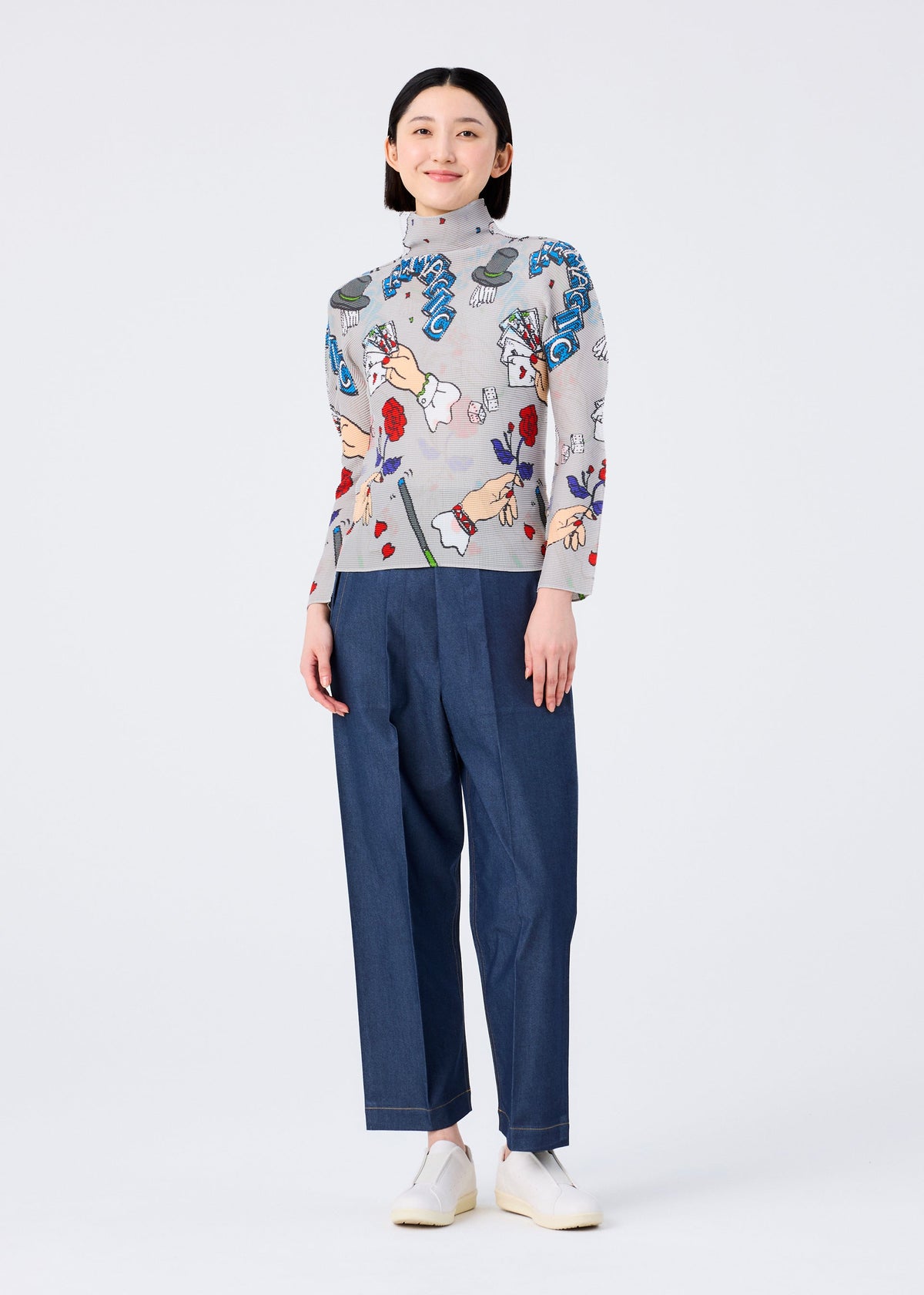 A person stands smiling in a MAGIC high-neck, long-sleeved top with colorful marker print by me ISSEY MIYAKE, blue high-waisted trousers, and white slip-on shoes against a plain white background. view 6 of 6