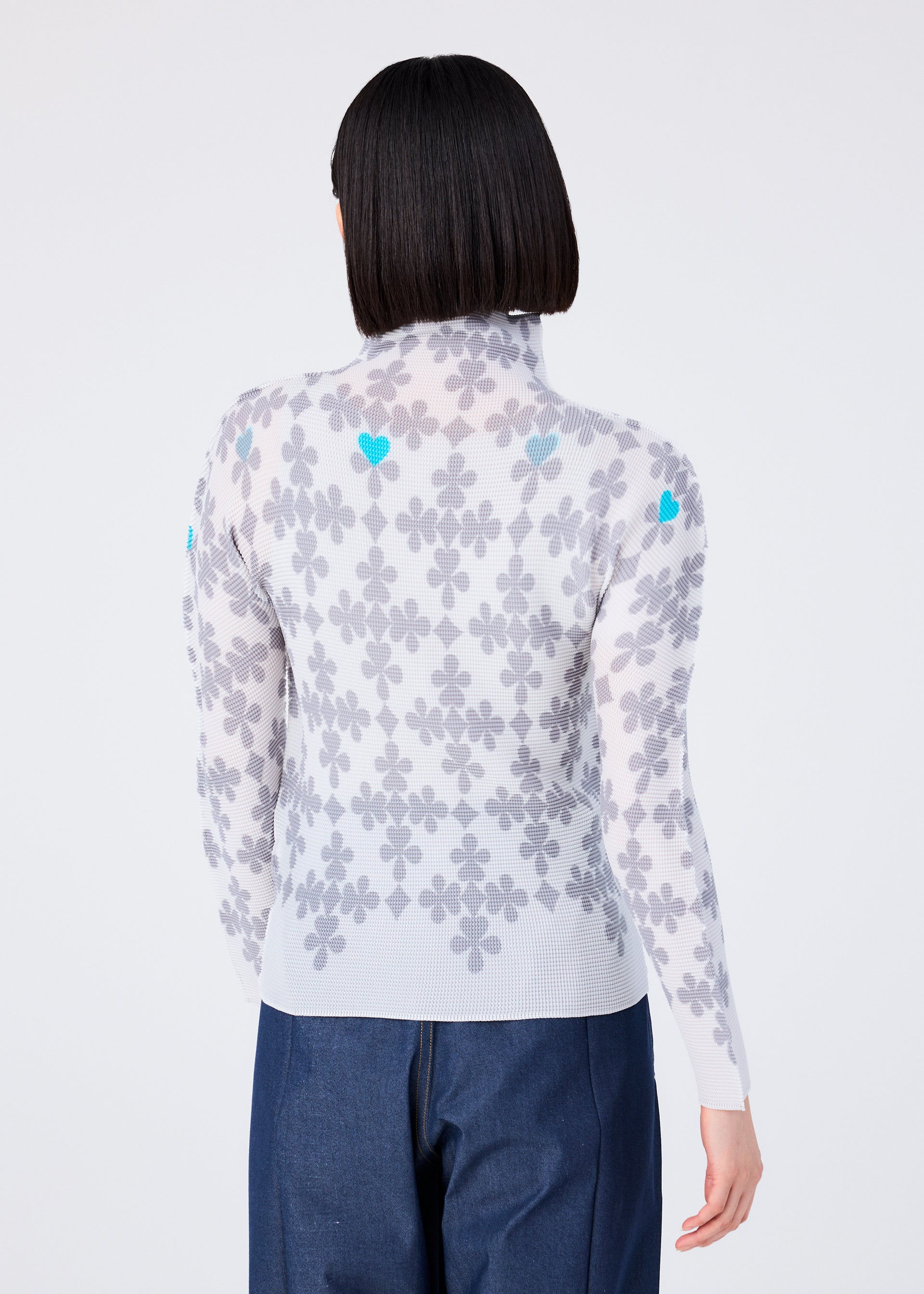 Zoomed view A person with short, straight black hair is seen from behind wearing the SUIT by me ISSEY MIYAKE—a high-neck, long-sleeved top with a gray floral pattern and blue accents, paired with dark blue pants—against a plain light background.