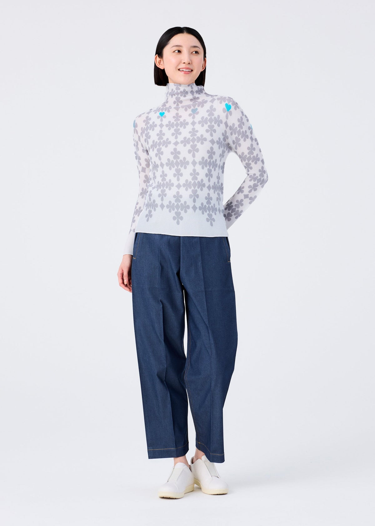 A woman smiles in a me ISSEY MIYAKE SUIT: a high-neck, long-sleeved top with a gray floral pattern, paired with blue wide-leg pants and white shoes, standing against a plain white background. view 7 of 7