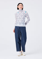 A woman smiles in a me ISSEY MIYAKE SUIT: a high-neck, long-sleeved top with a gray floral pattern, paired with blue wide-leg pants and white shoes, standing against a plain white background.