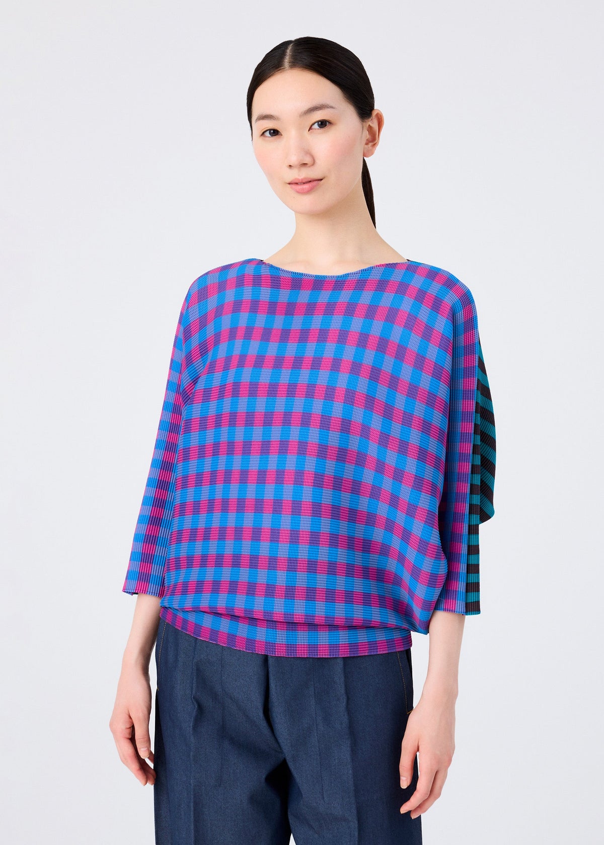 A woman with straight dark hair tied back wears the MAGIC STRIPE pleated blouse in blue and pink plaid with three-quarter sleeves by me ISSEY MIYAKE, paired with navy pants, standing against a plain light background. view 5 of 8