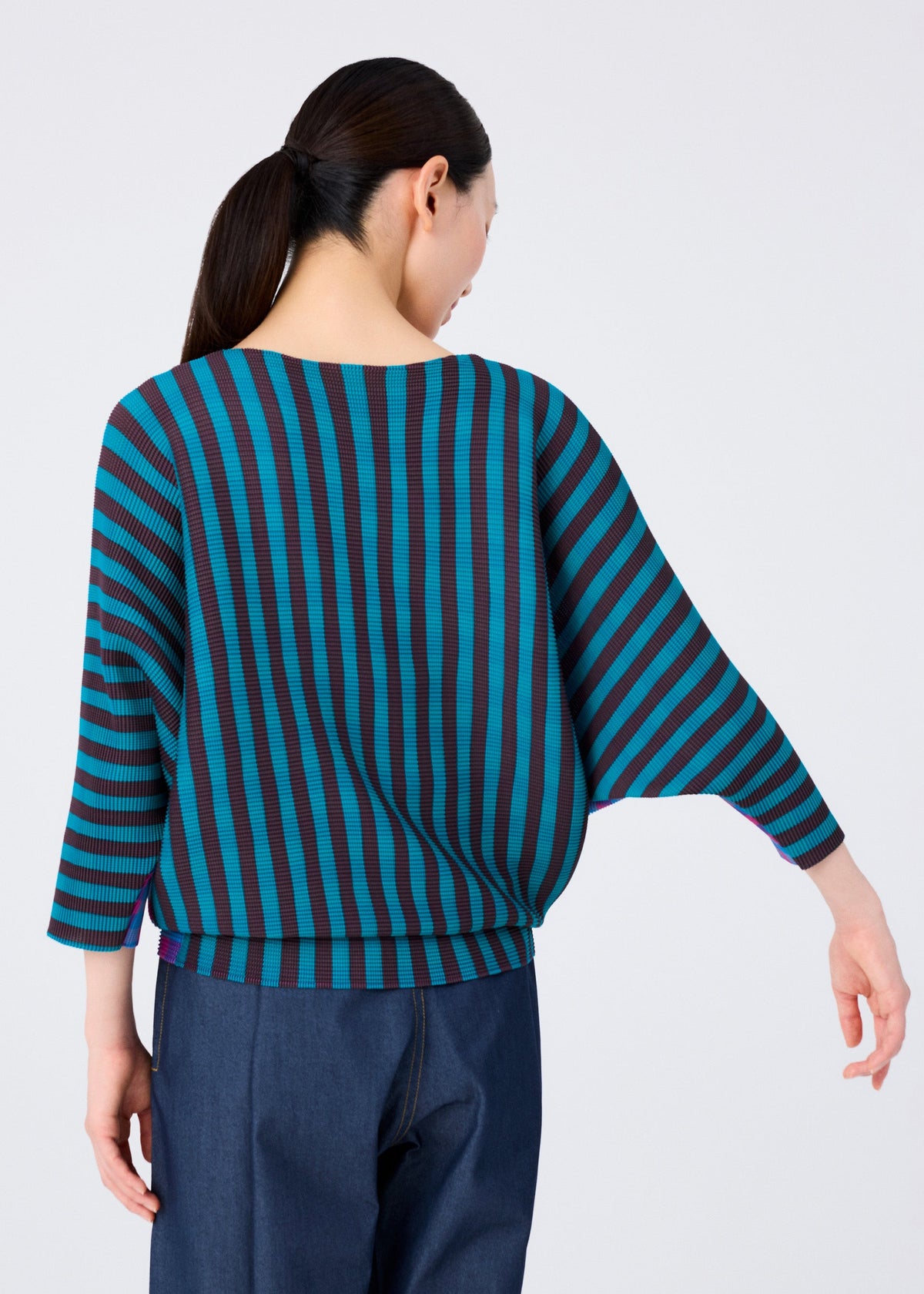 A woman with long dark hair in a low ponytail wears the MAGIC STRIPE top by me ISSEY MIYAKE—loose-fitting with wide sleeves and striped pattern—paired with dark pants, shown from the back against a plain light background. view 6 of 8