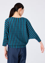 A woman with long dark hair in a low ponytail wears the MAGIC STRIPE top by me ISSEY MIYAKE—loose-fitting with wide sleeves and striped pattern—paired with dark pants, shown from the back against a plain light background.