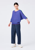 Against a white background, a person wears the me ISSEY MIYAKE MAGIC STRIPE blouse in blue and pink checks with wide-leg dark blue pants and white slip-on shoes, arms slightly outstretched and eyes cast downward.
