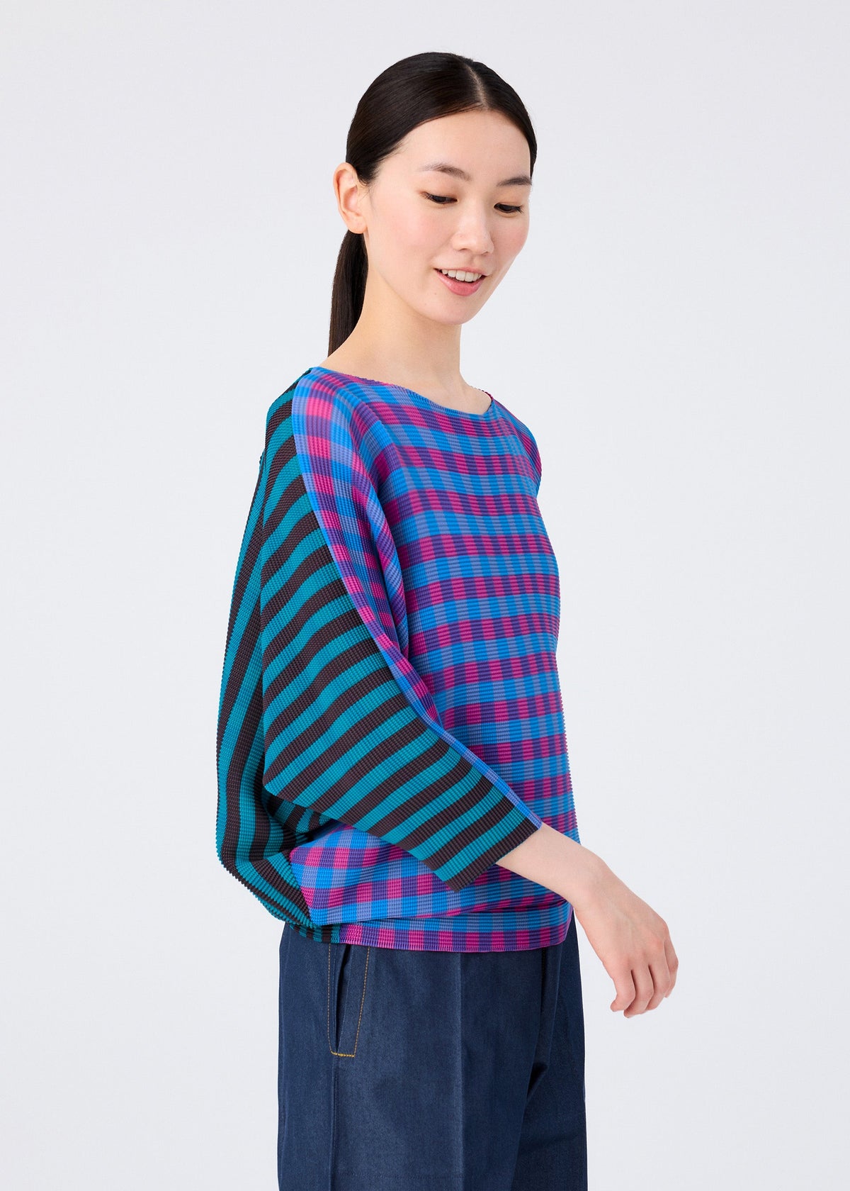 A woman with long dark hair tied back wears the MAGIC STRIPE top by me ISSEY MIYAKE, featuring blue and pink checkered and striped patterns, paired with dark blue pants, against a plain white background. view 8 of 8