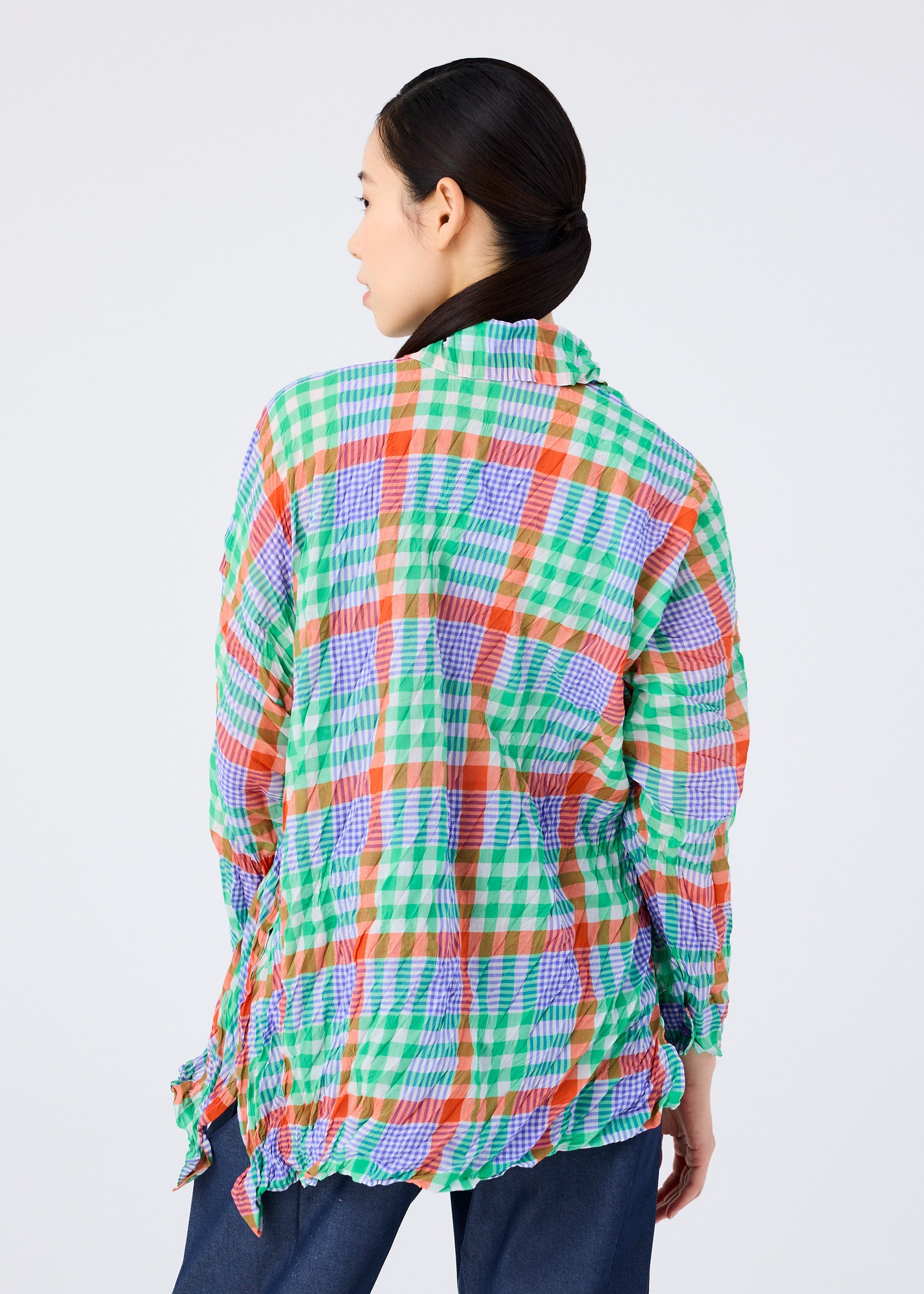 Zoomed view Seen from behind, a person with dark hair tied back wears the CHECK WHIPPED PLEATS SHIRT by me ISSEY MIYAKE, featuring green, orange, blue, and white yarn-dyed checks, paired with blue jeans against a plain light background.