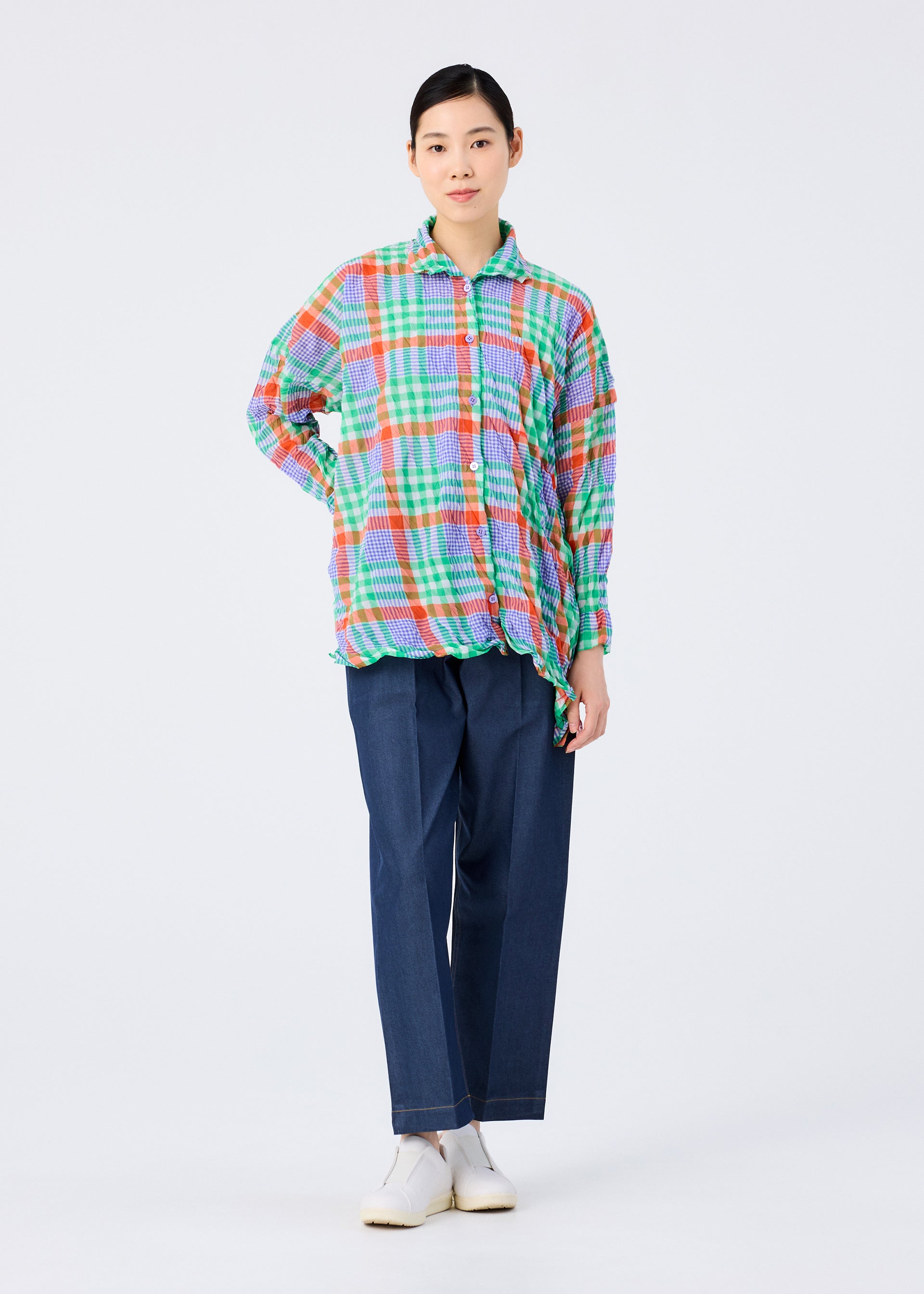 Zoomed view A person with dark hair tied back and a neutral expression wears the me ISSEY MIYAKE CHECK WHIPPED PLEATS SHIRT, featuring colorful yarn-dyed checks, paired with wide-leg blue pants and white shoes against a plain white background.