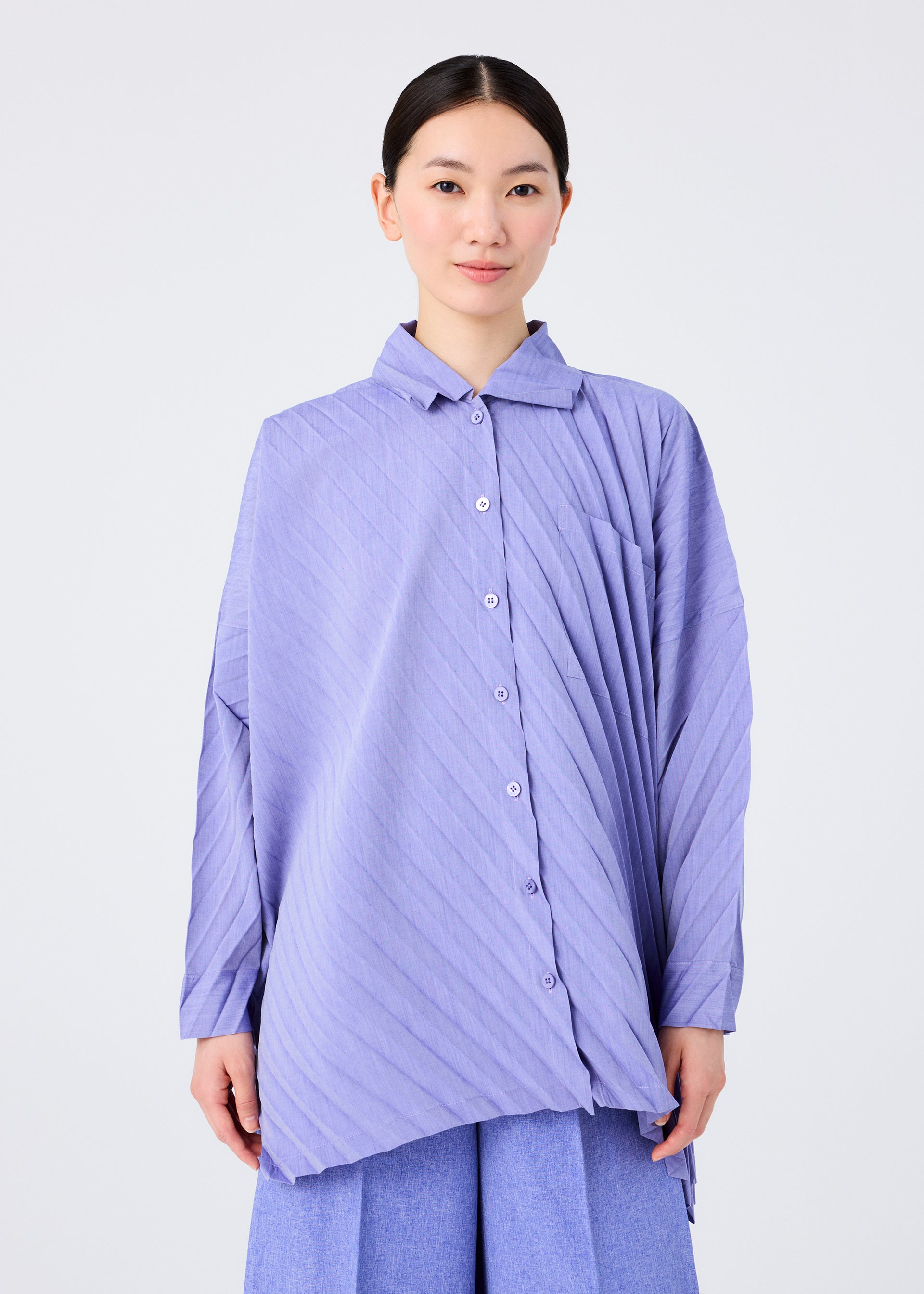 Zoomed view A person with dark hair tied back wears the ANGLE PLEATS SHIRT by me ISSEY MIYAKE, a lightweight, long-sleeved light purple top with diagonal stripes, paired with light blue pants against a plain light background.