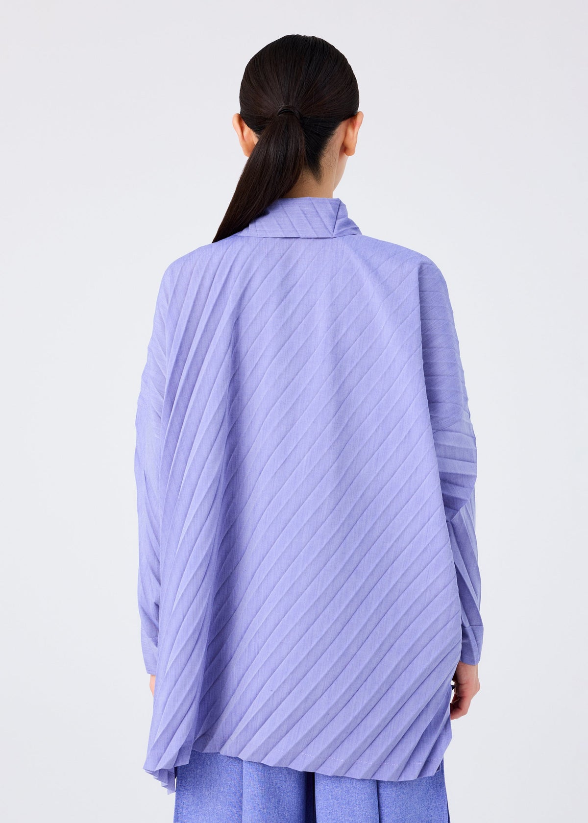Seen from behind, a person with long dark hair in a low ponytail wears the me ISSEY MIYAKE ANGLE PLEATS SHIRT in light purple with diagonal texture over a blue skirt, set against a plain white background. view 7 of 9