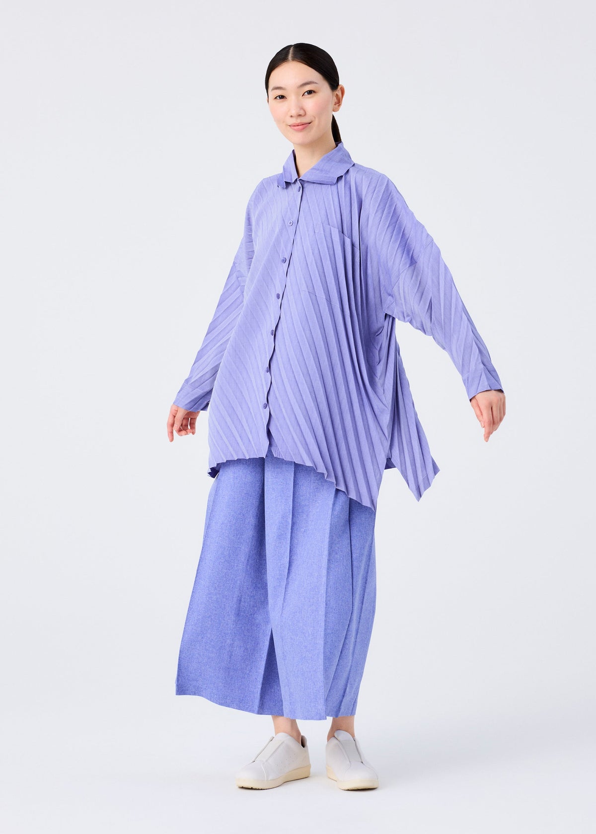 A person stands against a plain white background, smiling with arms slightly extended, wearing the me ISSEY MIYAKE ANGLE PLEATS SHIRT in lavender, styled with light blue wide-leg pants and white slip-on shoes. view 9 of 9