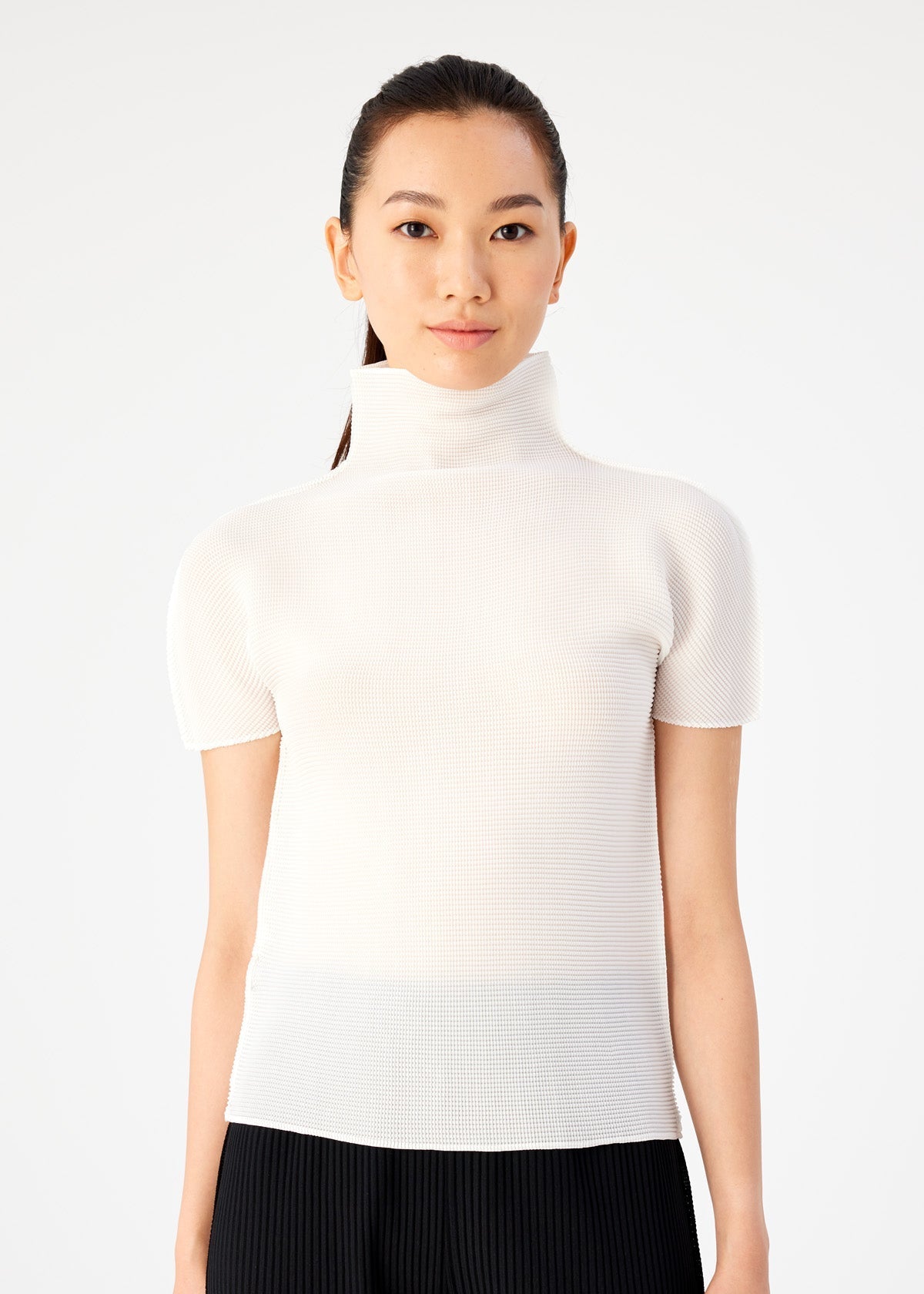 A woman with long dark hair pulled back wears me ISSEY MIYAKE STRETCH PLEATS black pants and a short-sleeved, high-neck top, standing against a plain white background and looking at the camera. view 4 of 6