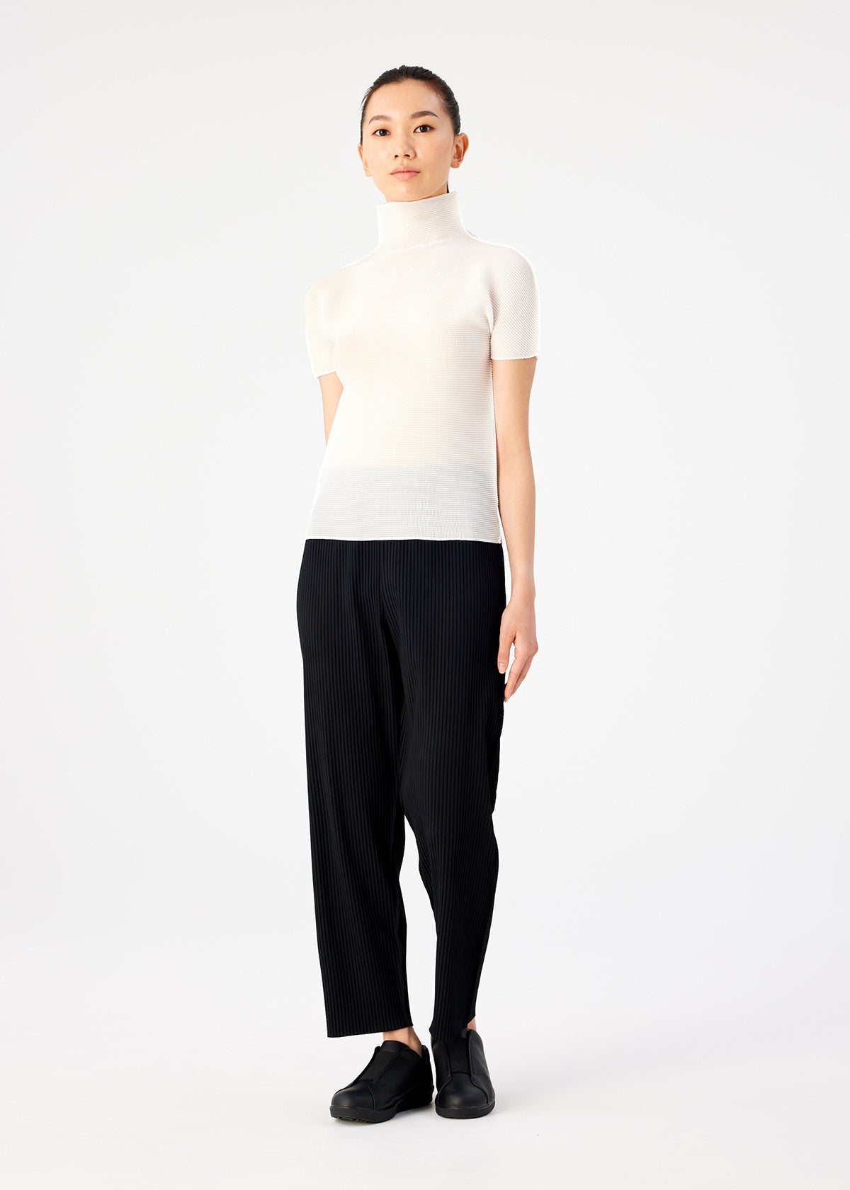 A woman stands against a white background wearing me ISSEY MIYAKE STRETCH PLEATS black pants, a short-sleeved high-neck top, and black slip-on shoes. She faces forward with a neutral expression and relaxed posture. view 6 of 6