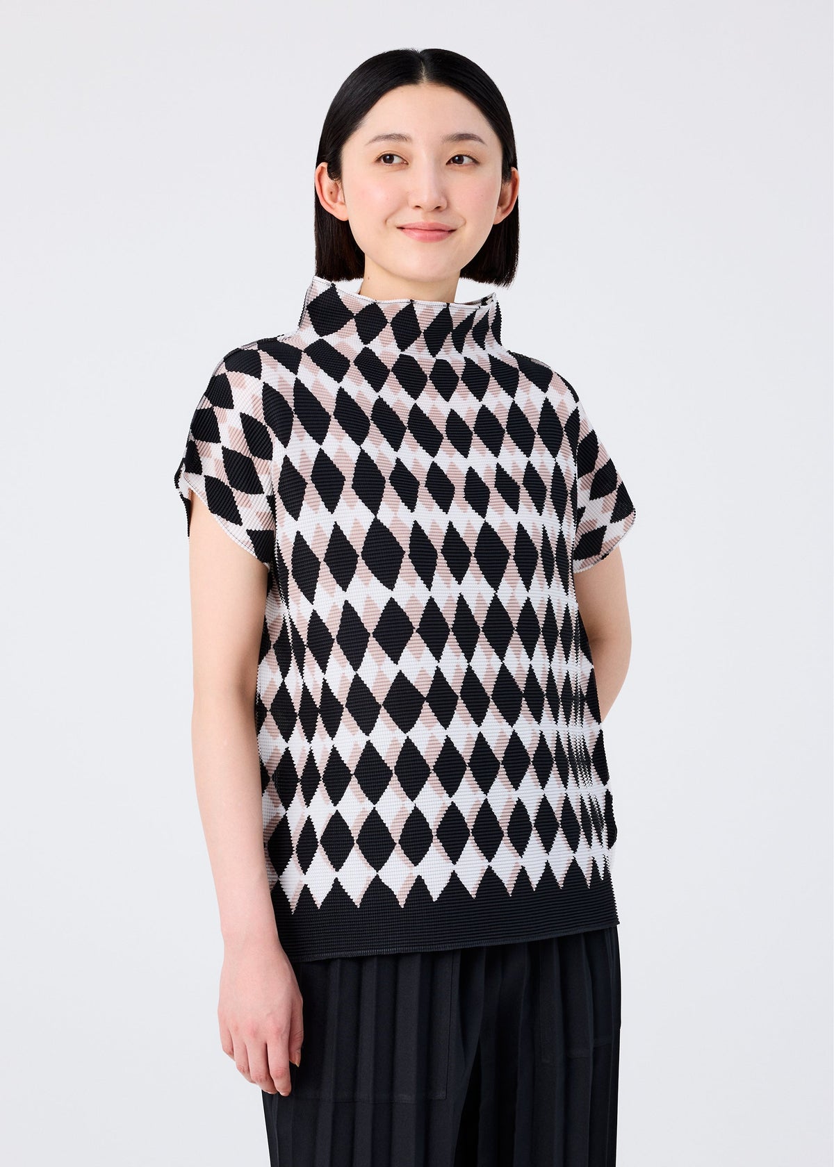 A woman with straight dark hair smiles and looks to the side, wearing the DIAMOND top by me ISSEY MIYAKE—a high-neck design with a black and white diamond motif—paired with black stretch pleated pants against a plain light background. view 4 of 6