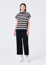 A person smiles against a white background, wearing the DIAMOND short-sleeve stretch pleats high-neck top with black and white diamond motif and black pleated wide-leg pants by me ISSEY MIYAKE, paired with white shoes.
