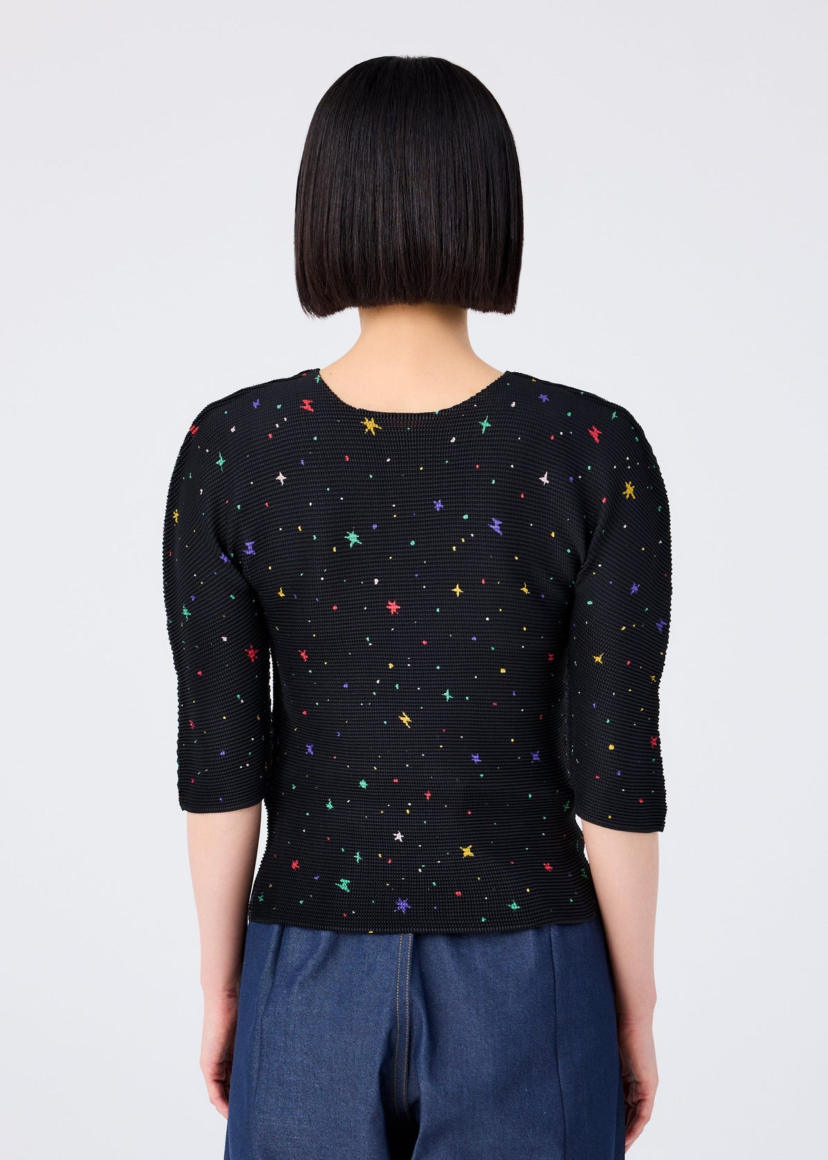 A person with straight, chin-length black hair is seen from behind wearing the TWINKLE STAR fitted round-neck short-sleeved top by me ISSEY MIYAKE, featuring colorful star patterns, paired with blue pants against a plain white background. view 5 of 6