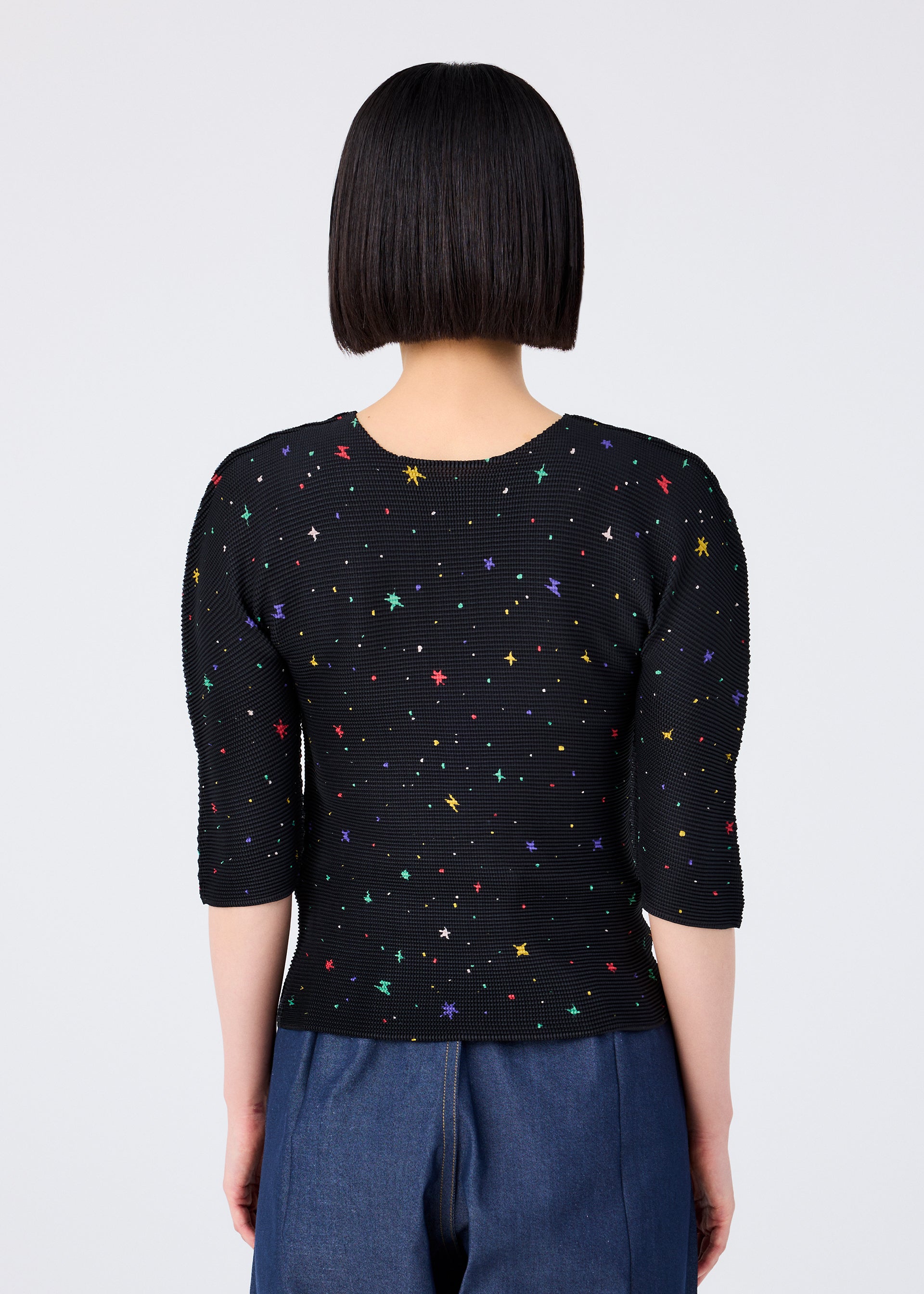 Zoomed view A person with straight, chin-length black hair is seen from behind wearing the TWINKLE STAR fitted round-neck short-sleeved top by me ISSEY MIYAKE, featuring colorful star patterns, paired with blue pants against a plain white background.