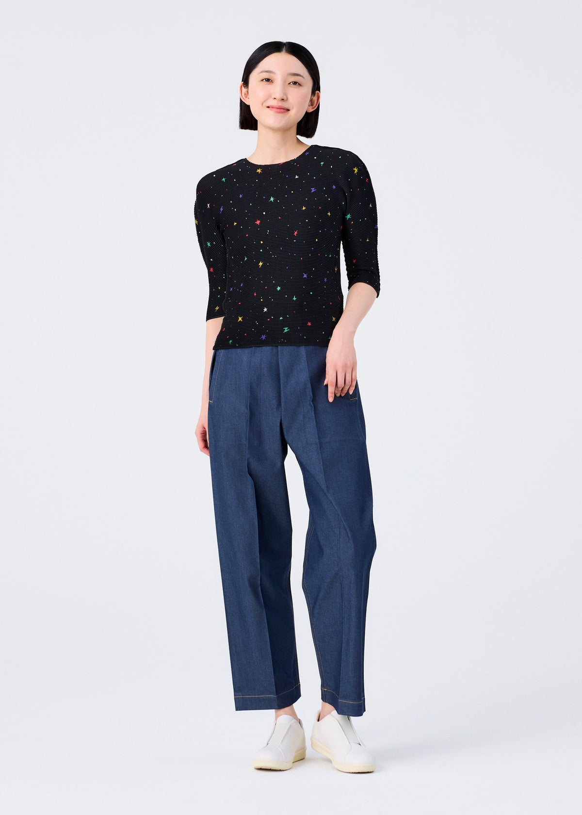 A person stands facing forward in the TWINKLE STAR round-neck top from me ISSEY MIYAKE, featuring a colorful print, paired with loose blue pants and white slip-on shoes, set against a plain white background. view 6 of 6