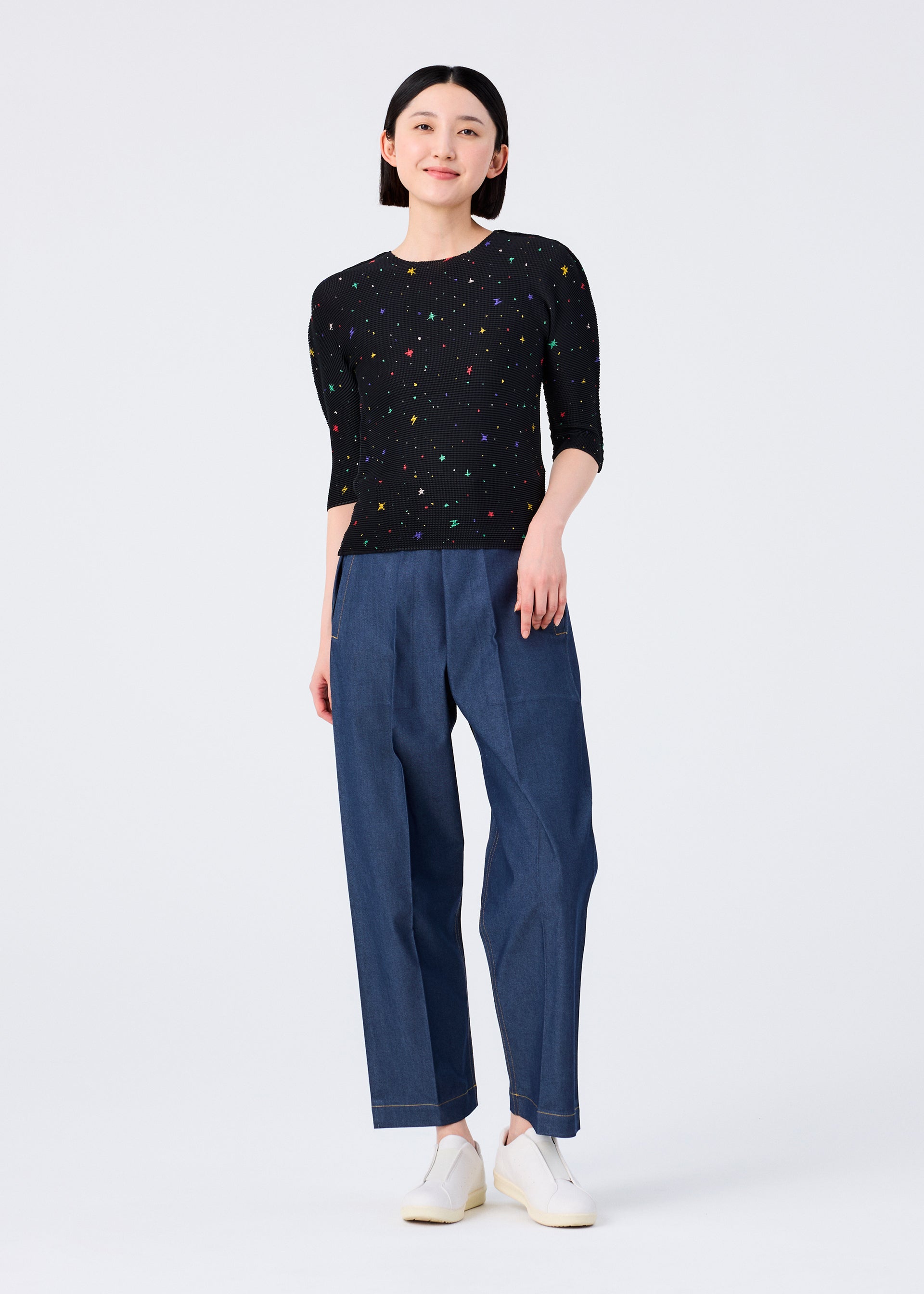 Zoomed view A person stands facing forward in the TWINKLE STAR round-neck top from me ISSEY MIYAKE, featuring a colorful print, paired with loose blue pants and white slip-on shoes, set against a plain white background.