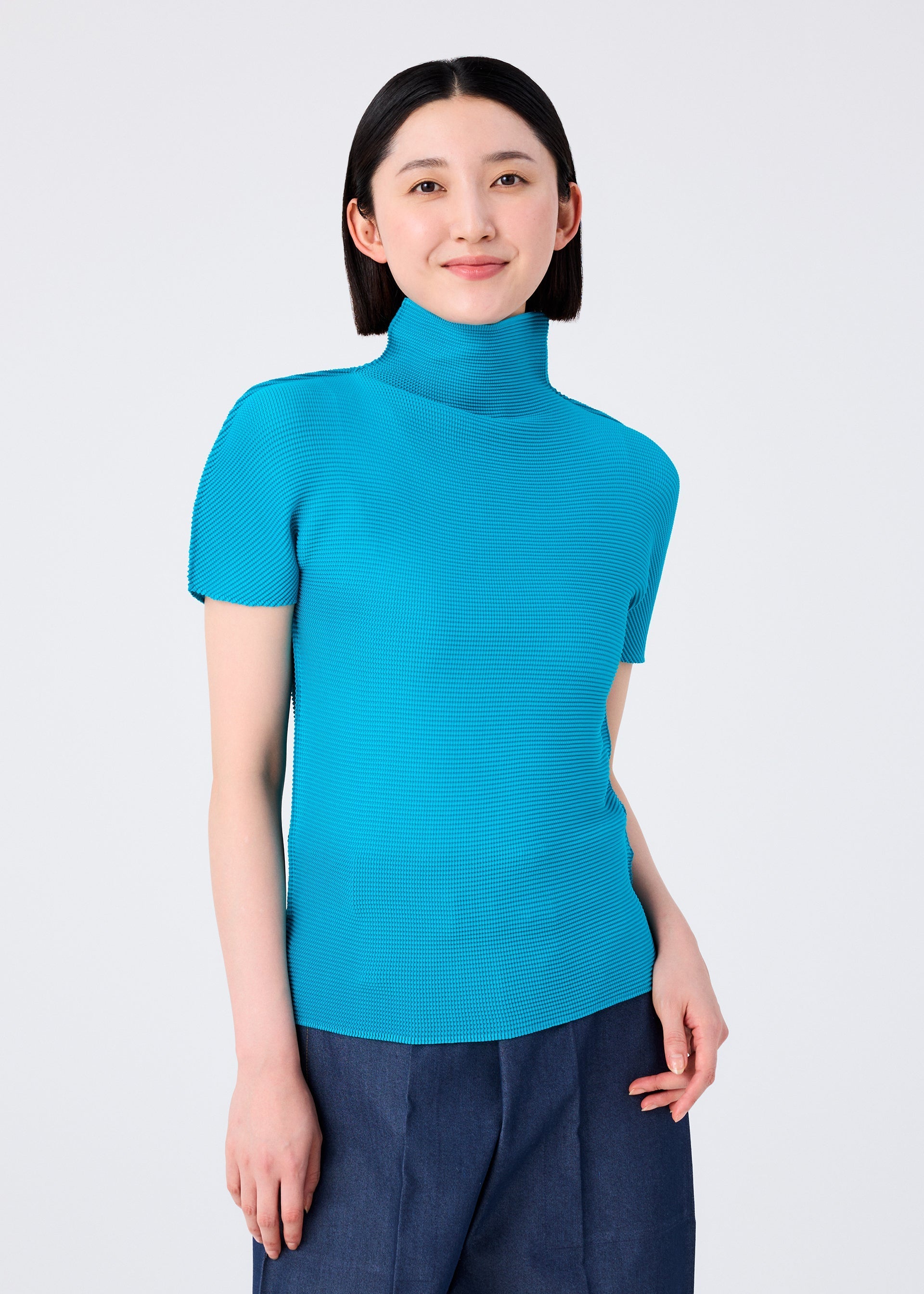 Zoomed view A person with straight black hair wears the STRETCH PLEATS 3 bright blue short-sleeved high-neck top by me ISSEY MIYAKE and dark pants, standing against a plain light background, smiling softly at the camera.