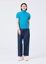 A woman faces forward, smiling, wearing the me ISSEY MIYAKE STRETCH PLEATS 3 bright blue short-sleeved high-neck top with loose dark blue pants and white shoes, set against a plain light background.