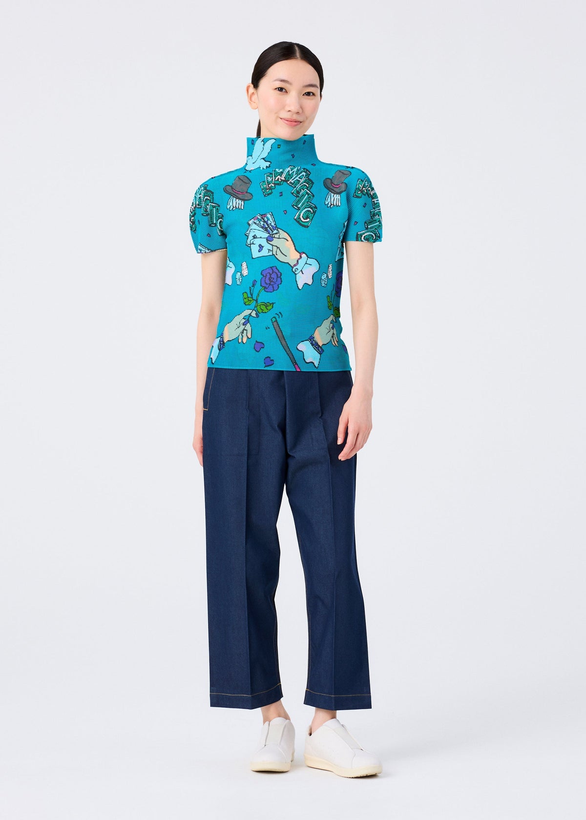 A woman stands against a white background wearing the MAGIC high-neck short-sleeved top by me ISSEY MIYAKE, featuring a turquoise magic marker print, paired with dark blue wide-leg pants and white slip-on shoes. She smiles slightly with her hands by her sides. view 6 of 6
