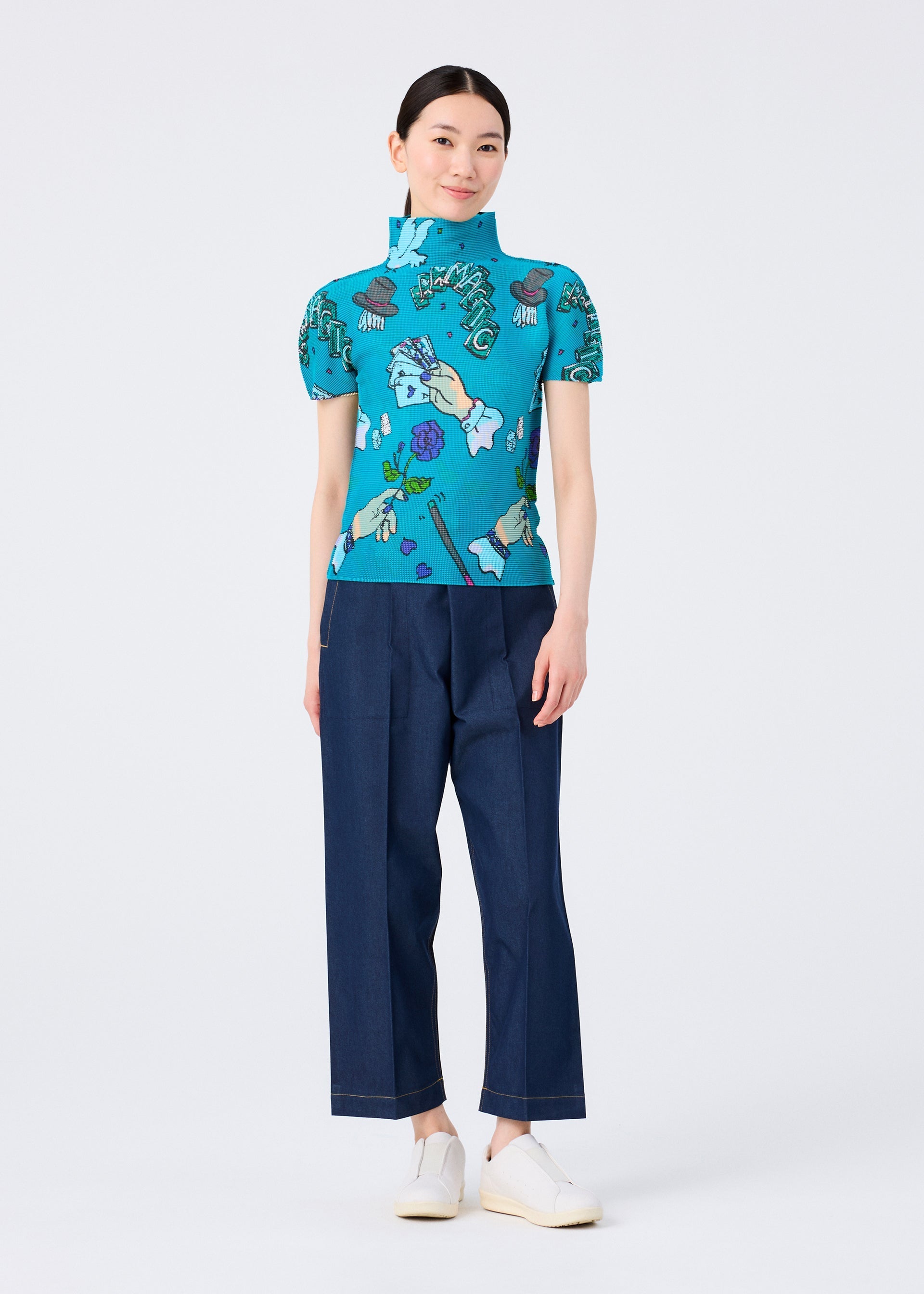 Zoomed view A woman stands against a white background wearing the MAGIC high-neck short-sleeved top by me ISSEY MIYAKE, featuring a turquoise magic marker print, paired with dark blue wide-leg pants and white slip-on shoes. She smiles slightly with her hands by her sides.