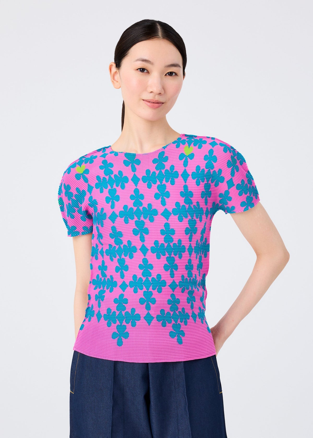 A woman with straight dark hair smiles subtly against a light background, wearing the SUIT by me ISSEY MIYAKE—a short-sleeved stretch pleats top with a blue floral pattern and dark blue pants—one hand resting on her hip. view 5 of 7