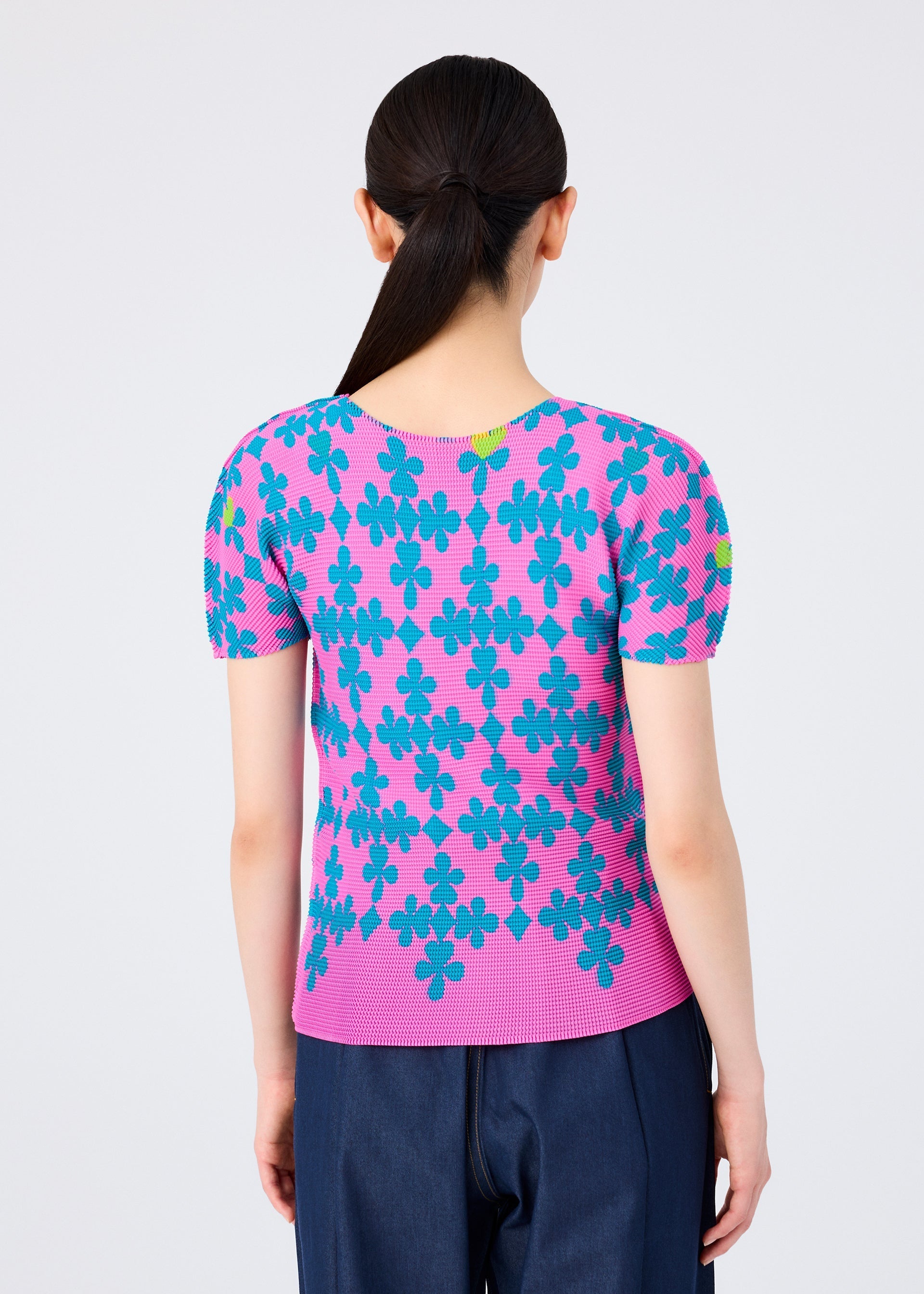 Zoomed view A person with long dark hair in a low ponytail is shown from the back, wearing the SUIT by me ISSEY MIYAKE—short-sleeved stretch pleats top with blue and green floral patterns and dark blue pants—against a plain light background.