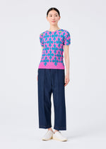 A woman stands against a plain background wearing the SUIT by me ISSEY MIYAKE: a pink, short-sleeved top with a blue playing card motif, wide-leg dark blue pants, and white slip-on shoes. Her hair is tied back as she glances to the side.