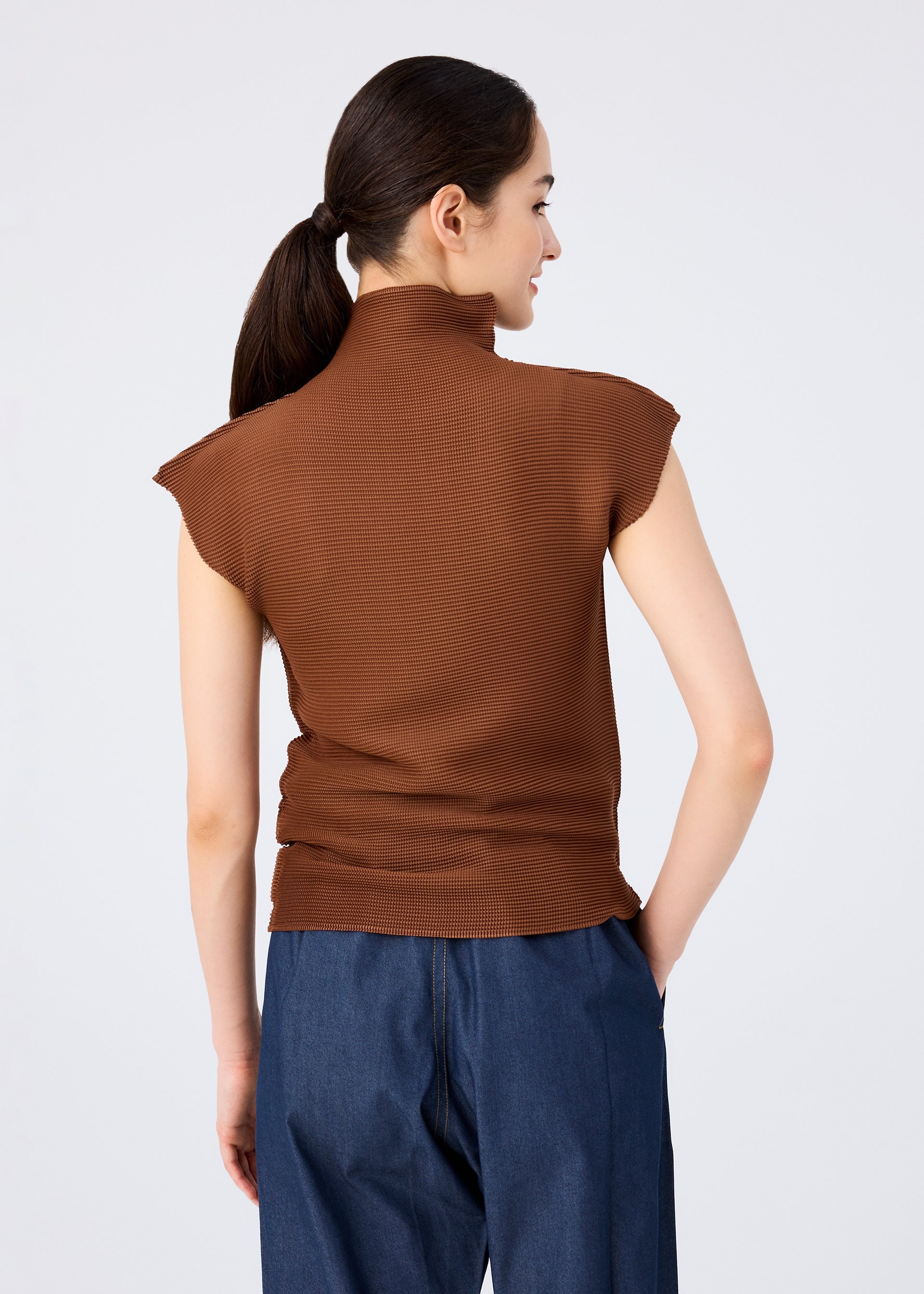 Zoomed view A woman with dark hair in a low ponytail is seen from behind, wearing the brown STRETCH PLEATS 4 high-neck French sleeve top by me ISSEY MIYAKE and blue denim pants against a light gray background.